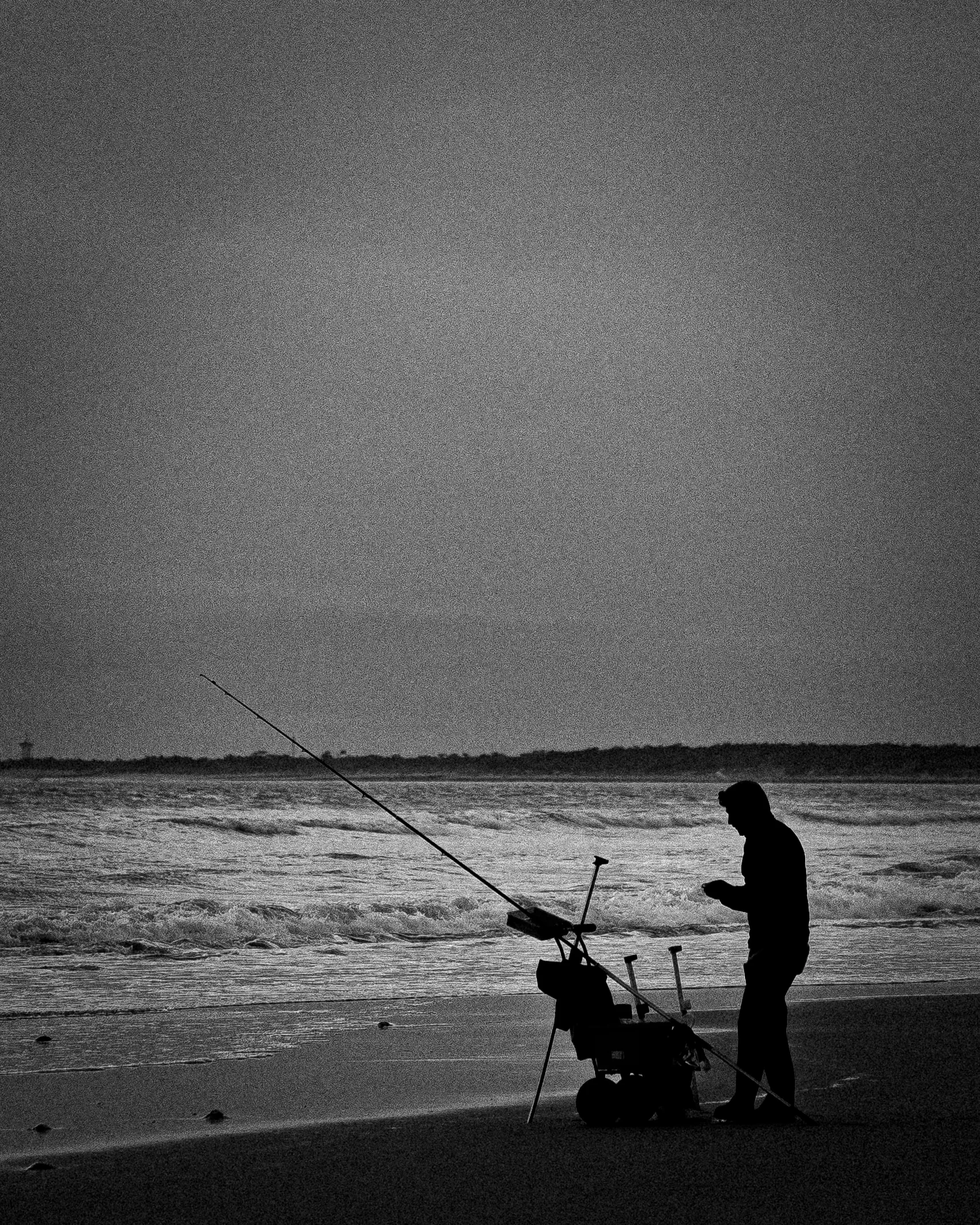 Silhouette d'un pêcheur debout sur la plage, avec une canne à pêche et un chariot, regardant son téléphone, face à la mer au coucher du soleil.