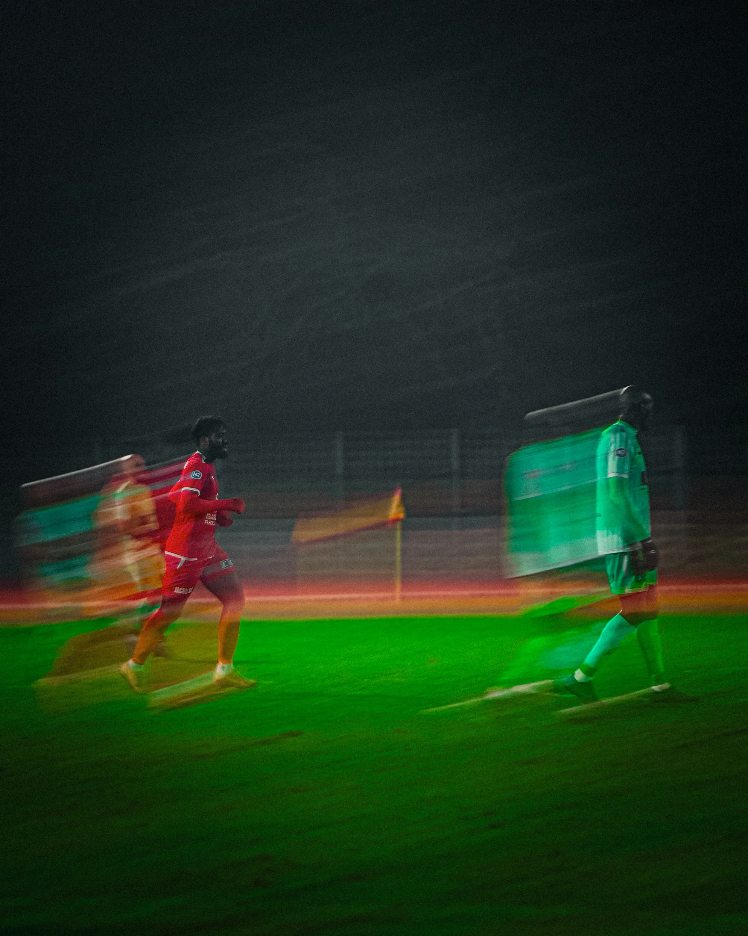 Trois joueurs courent sur un terrain de football la nuit, un des joueurs porte un maillot rouge, un autre porte une tenue verte avec des sacs de glace sur la tête.
