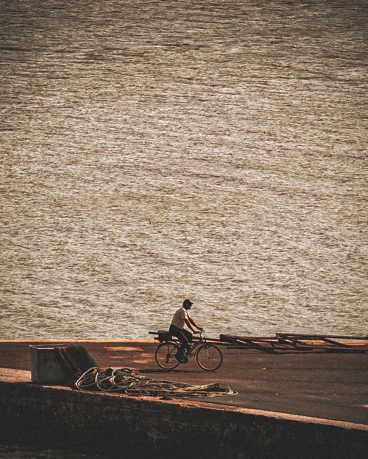 Un homme pédalant à vélo sur un chemin proche de l'eau, avec des cordes enroulées à côté. Le paysage est calme et le soleil semble se coucher ou se lever.