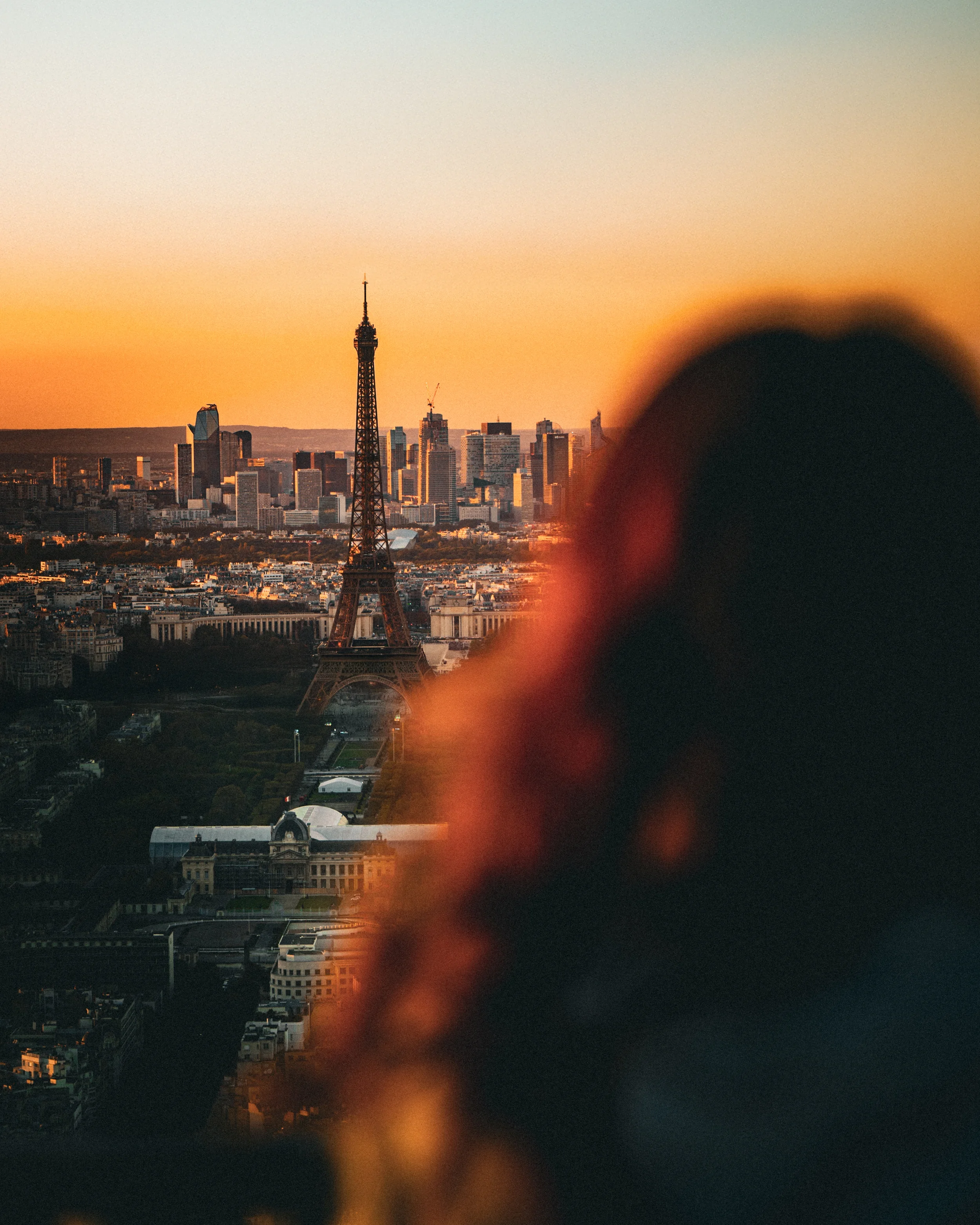 Une vue de Paris au coucher du soleil avec la Tour Eiffel en centre, un reflet flou en premier plan, et le skyline moderne en arrière-plan.