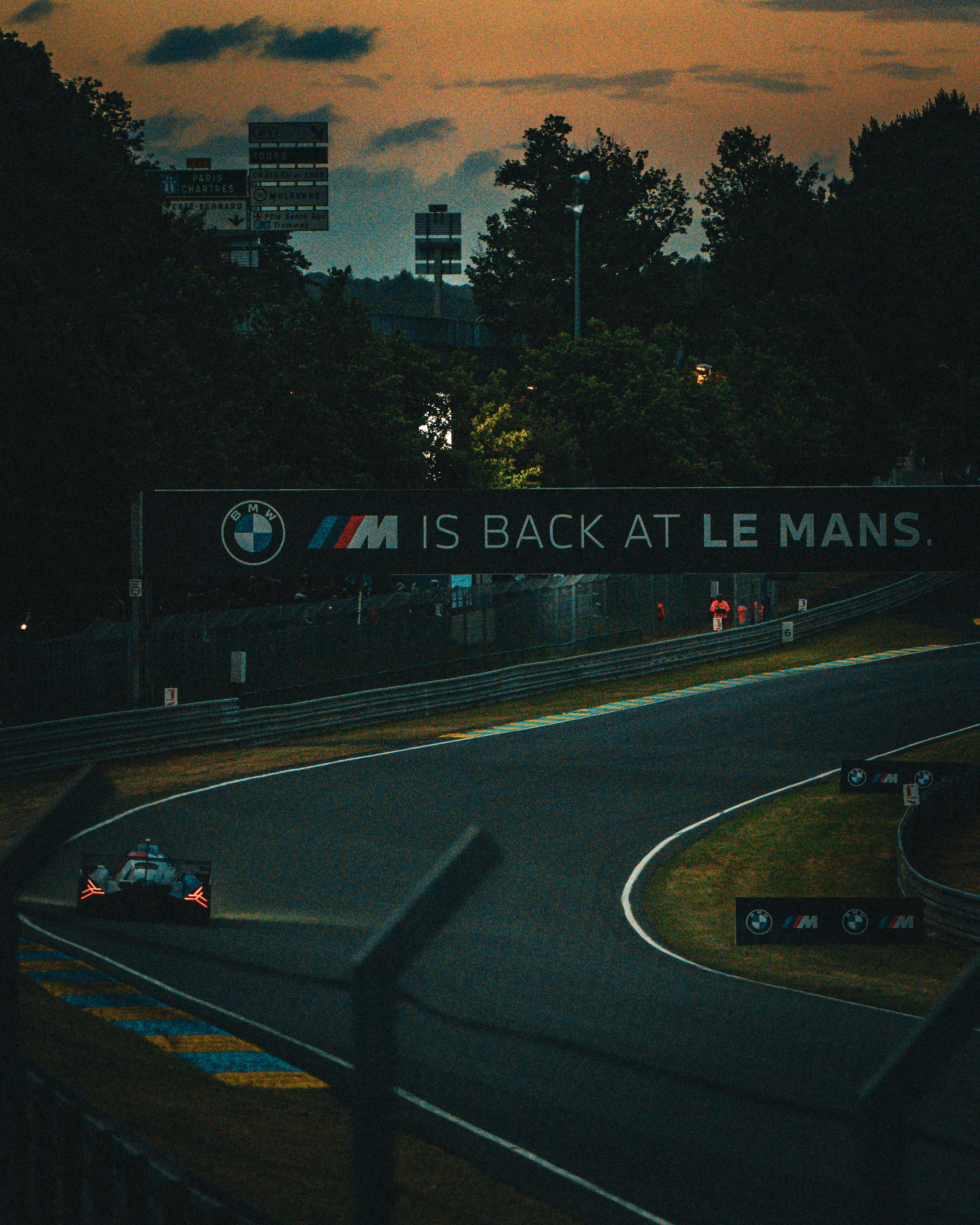 Une voiture de course sur un circuit lors du Grand Prix de France, avec un panneau d'affichage annonçant le retour de BMW M au Mans, au coucher du soleil.
