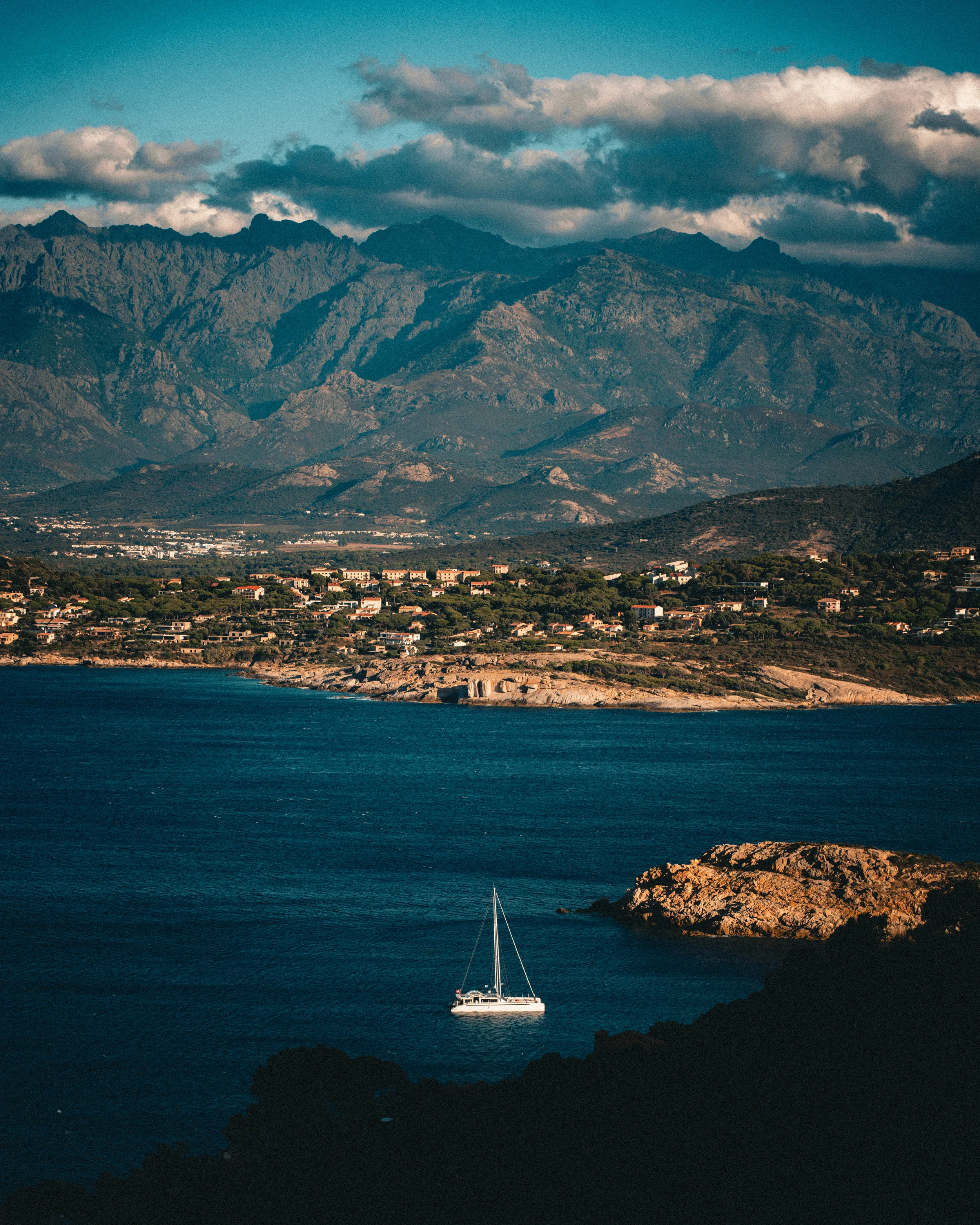 Paysage côtier avec mer, un voilier, une péninsule rocheuse et une ville en arrière-plan avec montagnes et ciel nuageux.