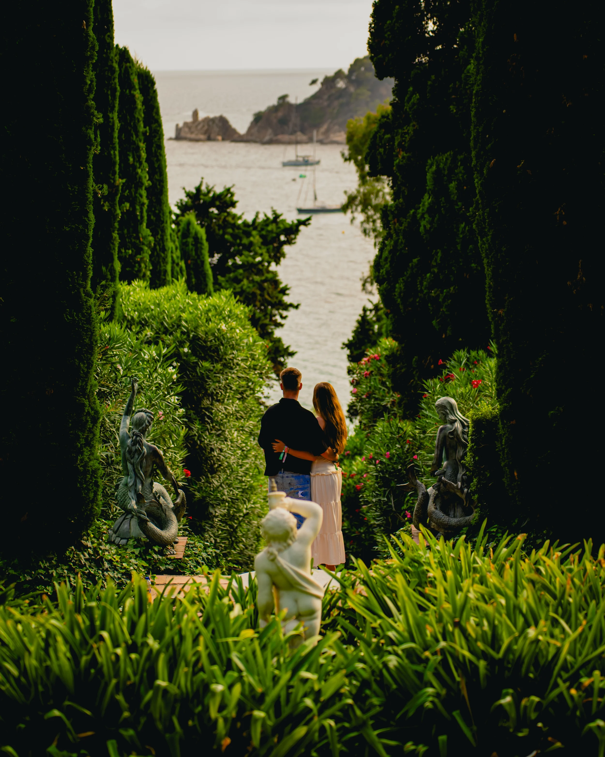 Un couple regarde vers la mer dans un jardin luxuriant entouré de statues en pierre, avec des arbres, des haies, et des fleurs, lors d'une journée ensoleillée.