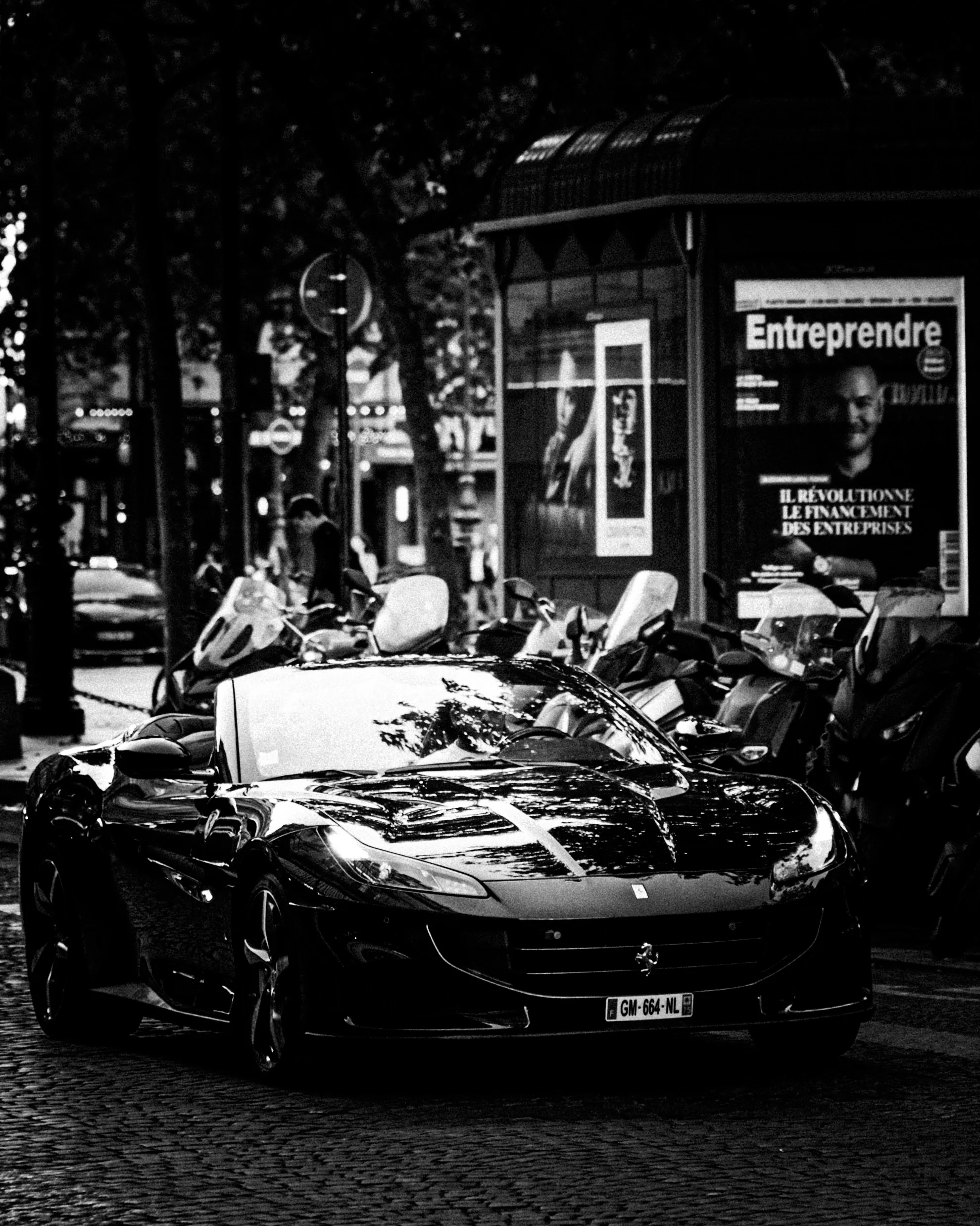 Une voiture de luxe noire stationnée dans une rue urbaine en soirée, avec des motos et des panneaux publicitaires autour.