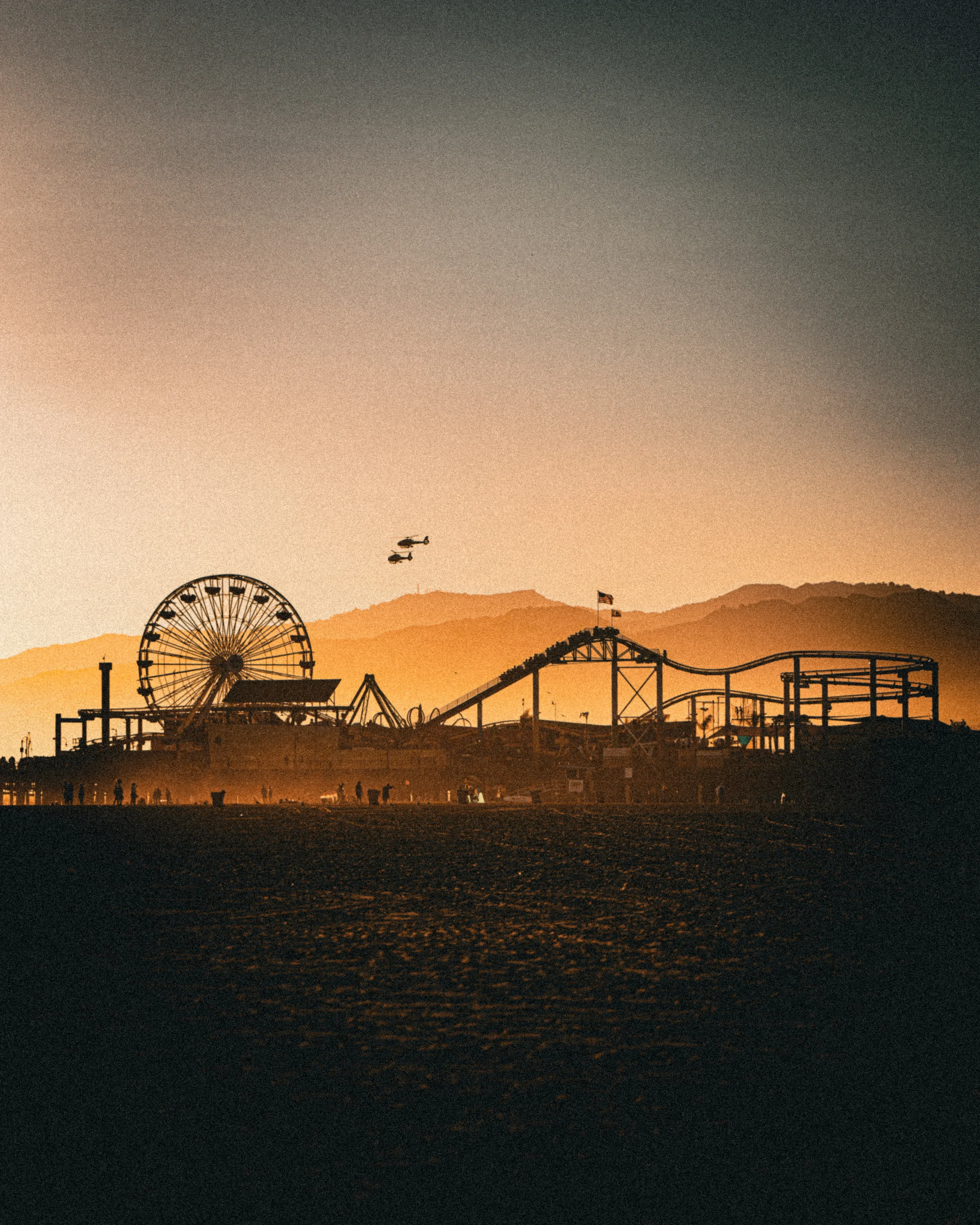 Vue d'une fête foraine au coucher du soleil avec une grande roue, des montagnes en arrière-plan, et des hélicoptères dans le ciel.