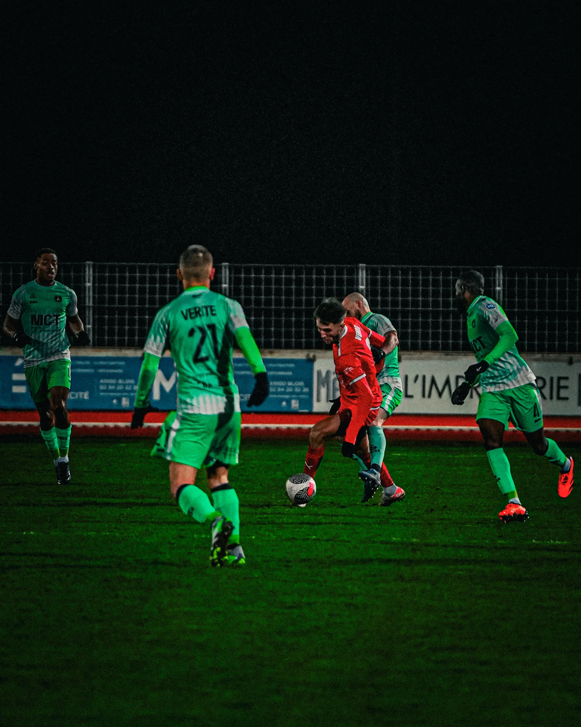 Des joueurs de football en train de jouer sur un terrain en nocturne, avec un joueur en rouge contrôlant le ballon entouré de joueurs en vert.