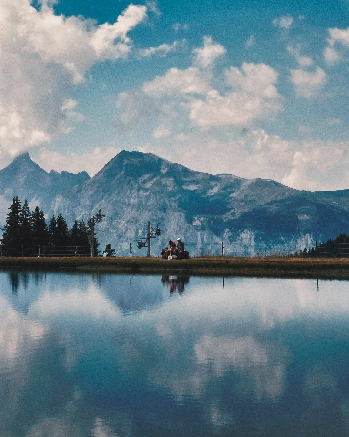 Montagnes majestueuses en arrière-plan avec un ciel partiellement nuageux, un lac calme en premier plan qui reflète le paysage, et deux personnes assises sur un banc au bord du lac, profitant de la vue.