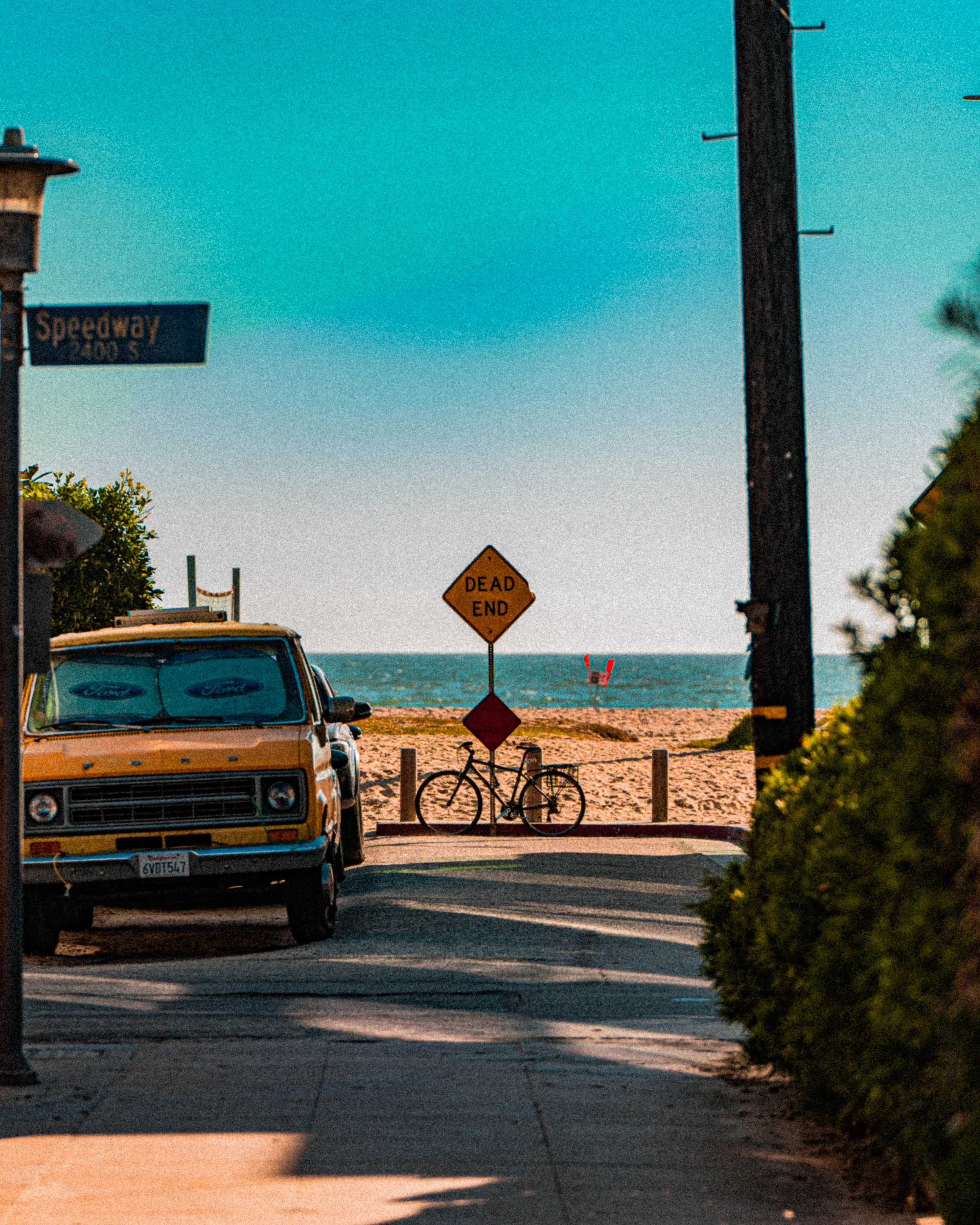 Une rue menant à la plage, avec une voiture jaune stationnée à gauche, un vélo posé près de l'entrée à droite, un panneau 'Dead End' au centre, à l'arrière la plage avec des dunes et la mer, et un lampadaire sur la gauche.