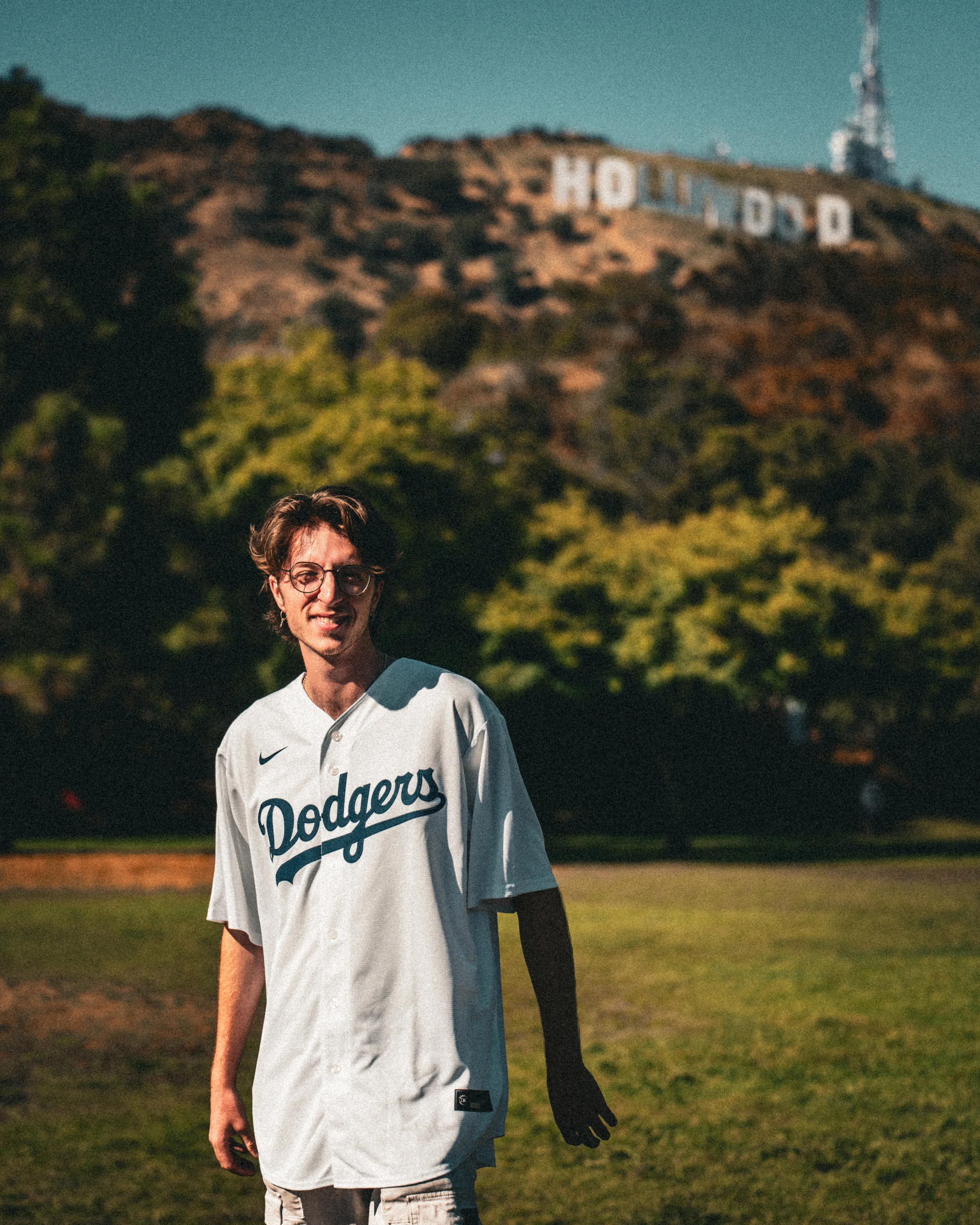 Un jeune homme souriant portant un maillot de baseball des Dodgers, se tenant dans un parc avec les collines de Hollywood en arrière-plan, incluant le célèbre panneau Hollywood.