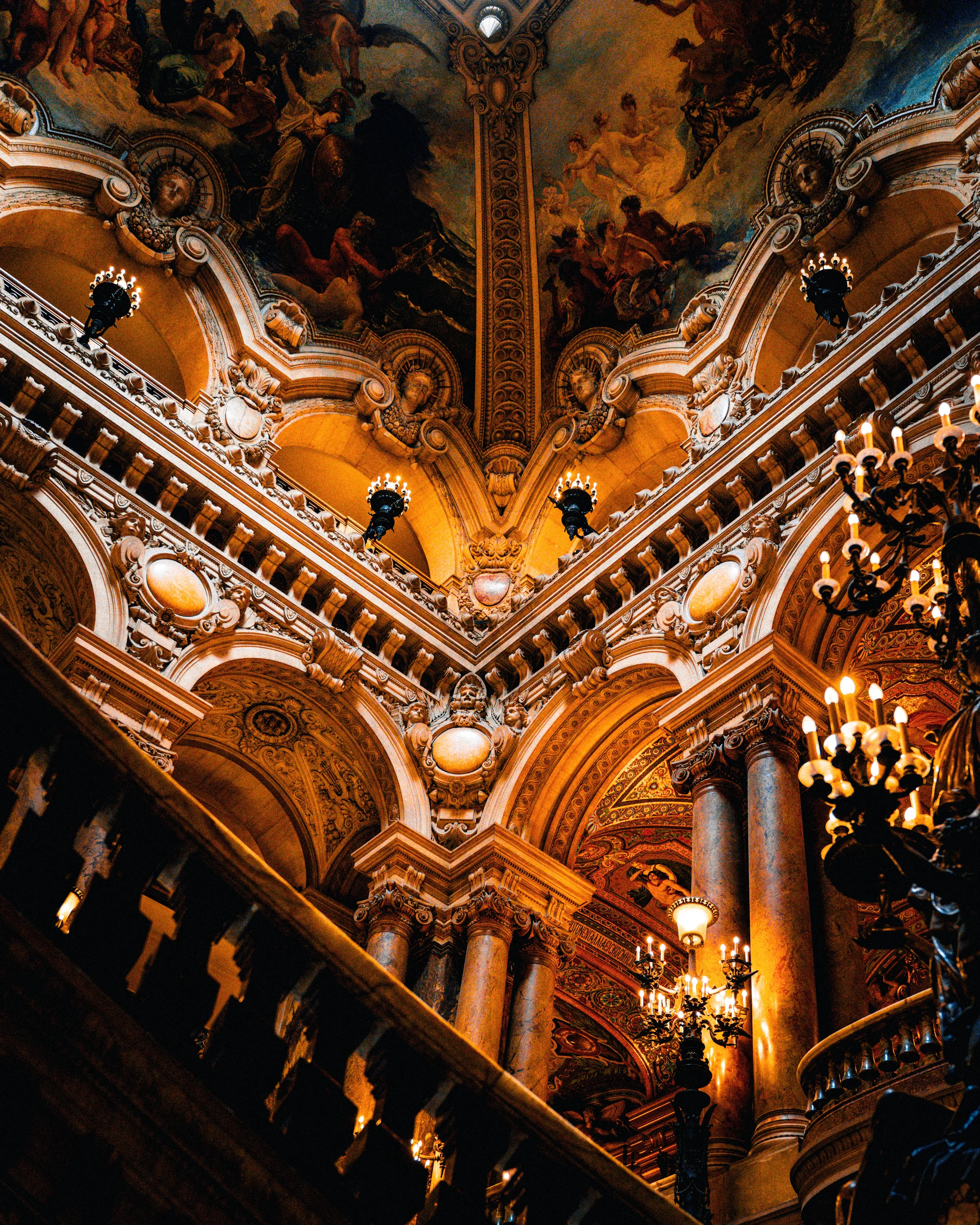 Intérieur somptueux d'un bâtiment historique avec des colonnes en marbre, des chandeliers en fer forgé, une fresque au plafond représentant des scènes mythologiques ou religieuses, et une décoration riche et détaillée.