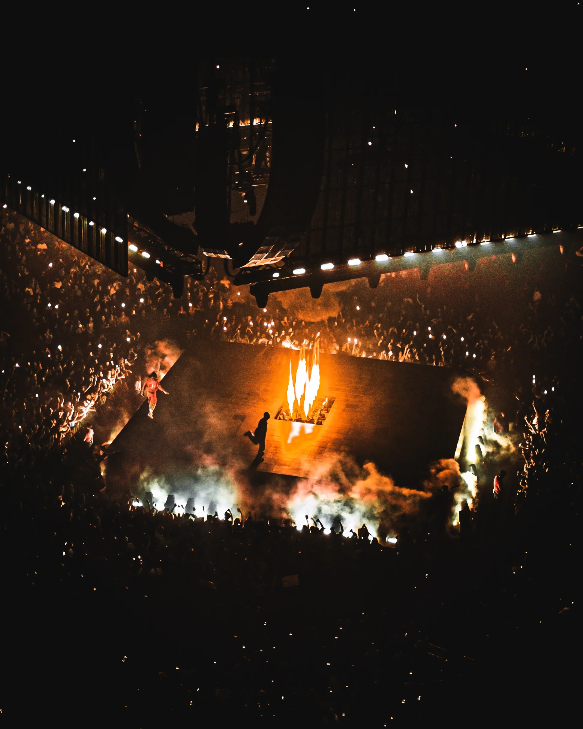 Scène de concert en plein air avec un grand feu au centre, entouré d'une foule nombreuse, illuminée par des lumières et une atmosphère de fête nocturne.