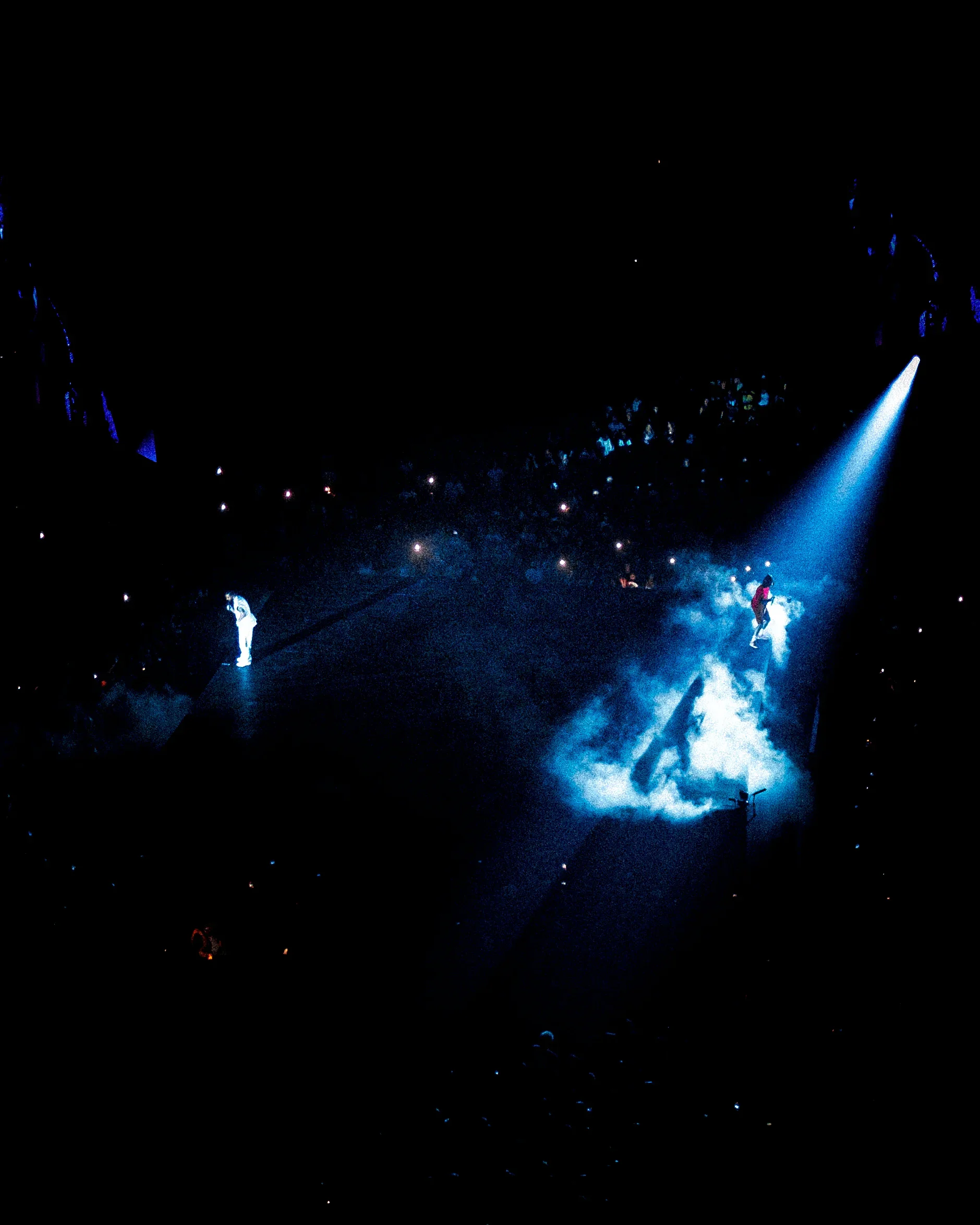 Une scène de concert avec deux artistes sur scène, éclairés par des projecteurs bleus, dans une ambiance sombre avec des effets de fumée, et un public avec des lumières.
