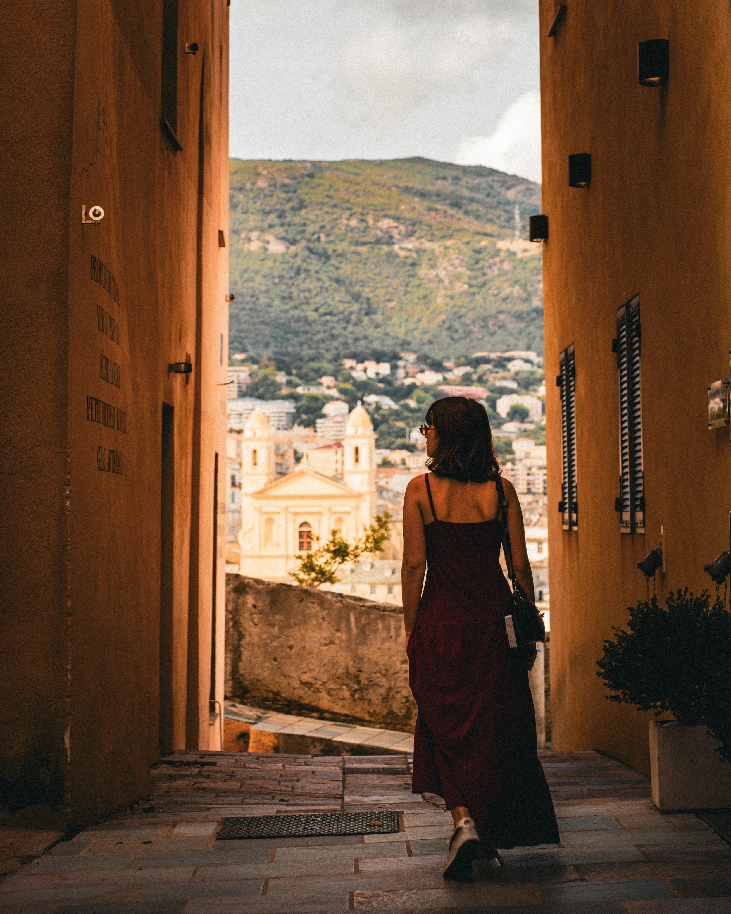 Une femme portant une robe rouge marche dans une allée entre deux bâtiments vers une vue sur une ville et une colline au loin.