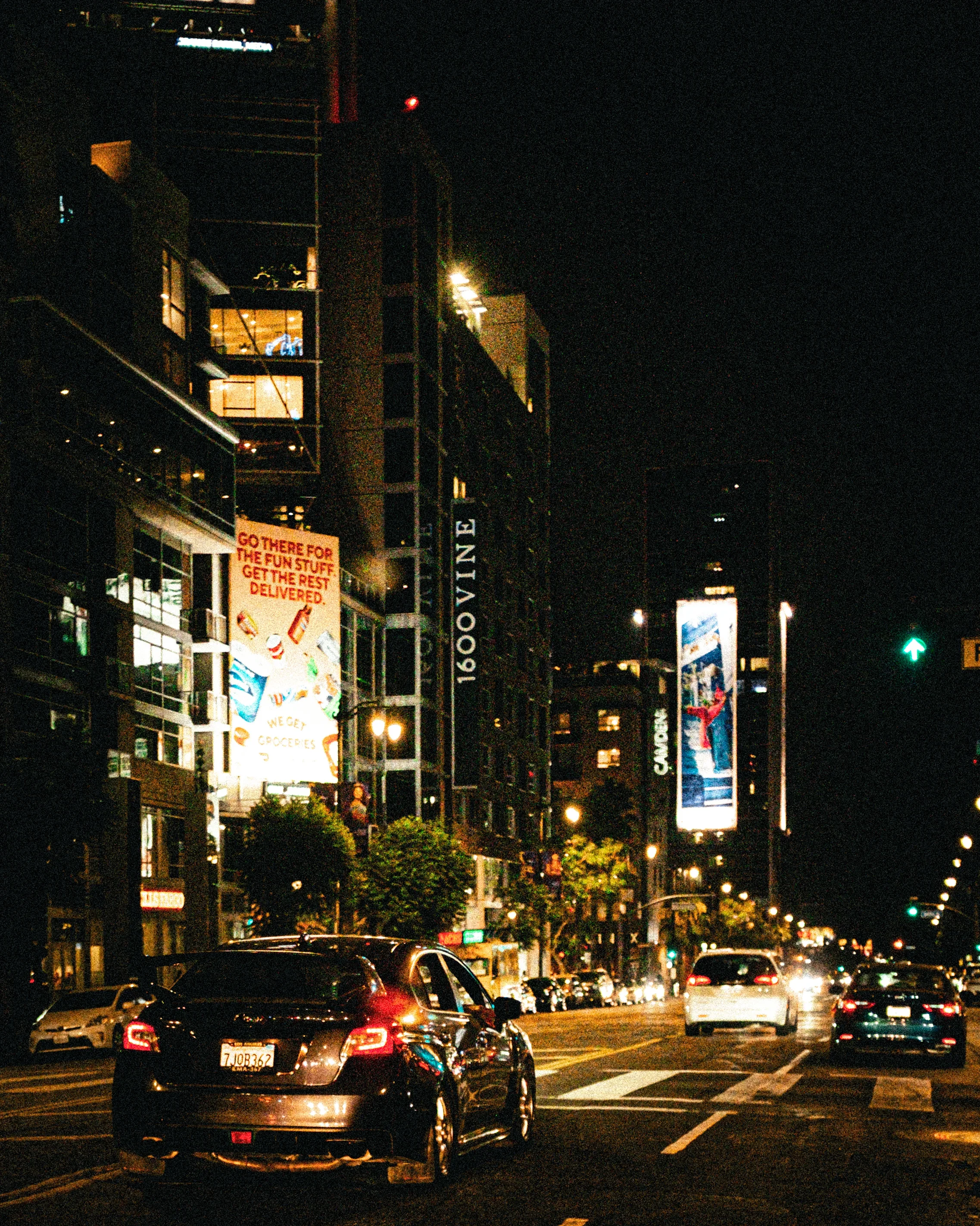 Une rue urbaine la nuit avec des bâtiments éclairés, des voitures stationnées ou passant, et des affiches publicitaires sur les façades.
