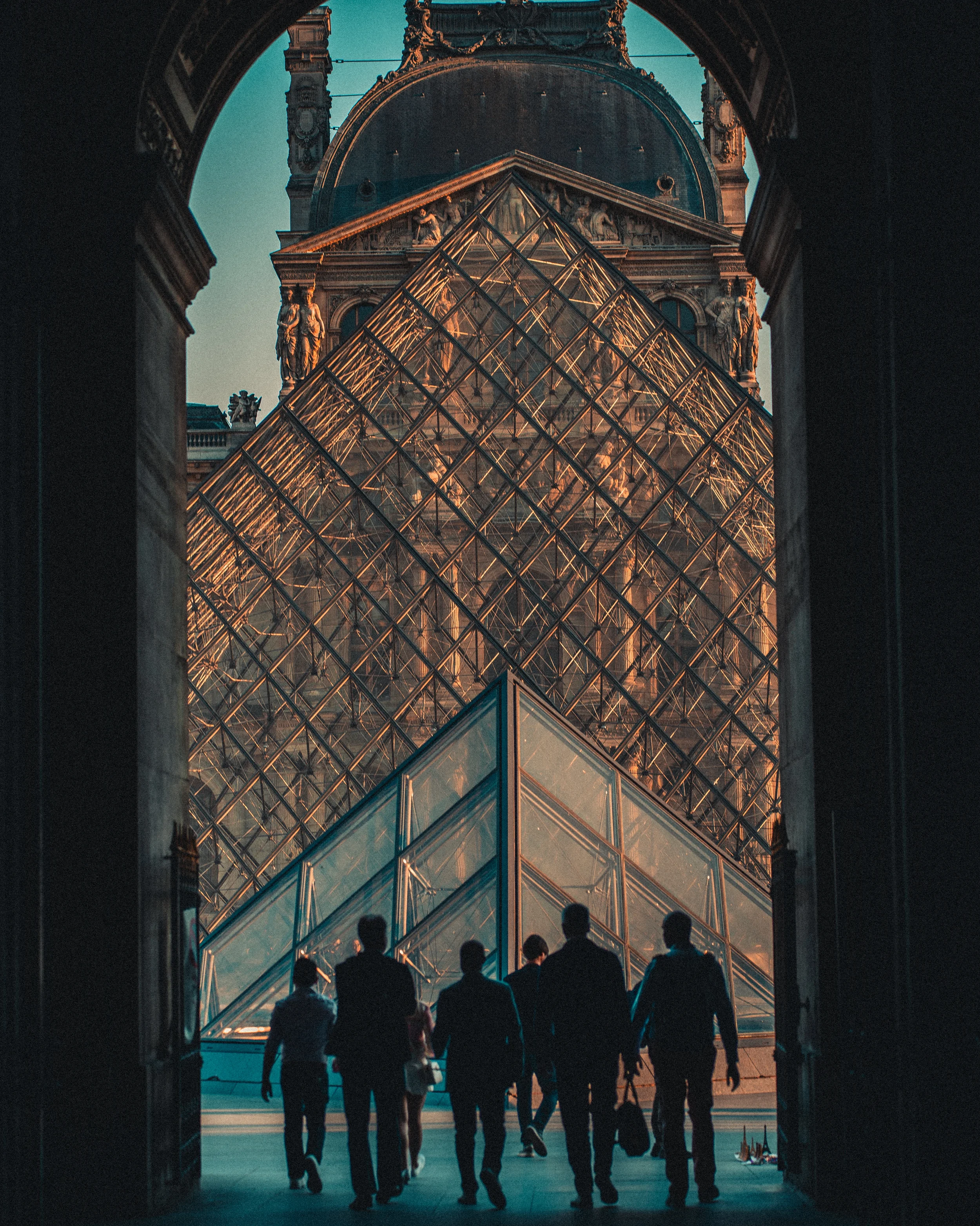 Groupe de personnes en costume marchant vers le musée du Louvre, avec la pyramide en verre emblématique en premier plan au coucher du soleil.