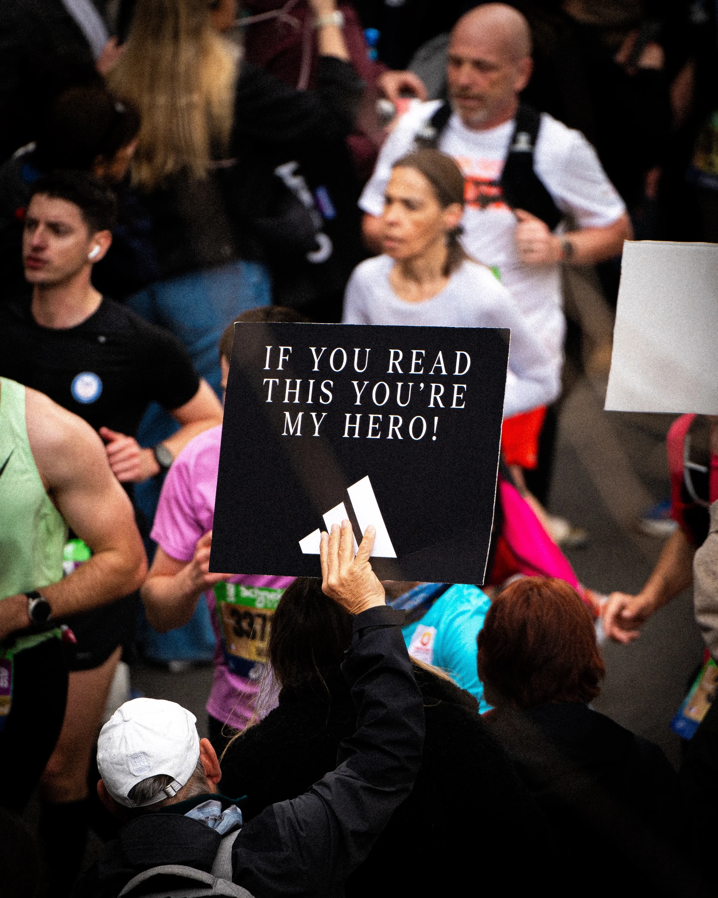 Une personne tient une pancarte noire avec l'inscription 'SI TU LIS CECI TU ES MON HÉROS !' lors d'une course ou d'une manifestation, entourée de plusieurs autres personnes.