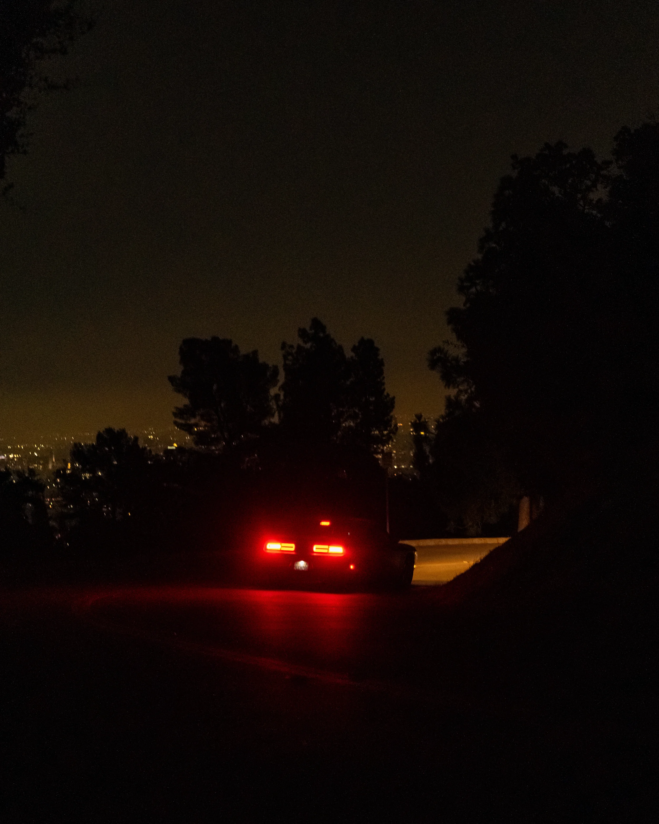 voiture noire avec feux arrière allumés sur une route sombre la nuit, entourée d'arbres et de silhouettes de villes à l'horizon