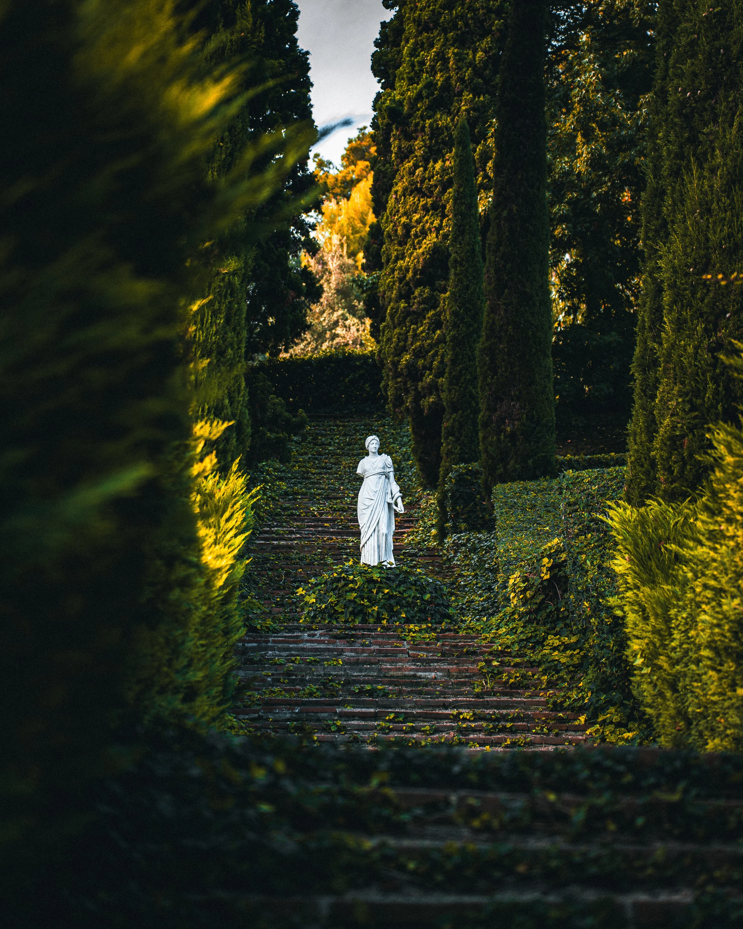 Statue blanche d'une femme dans un jardin avec des grands arbres.