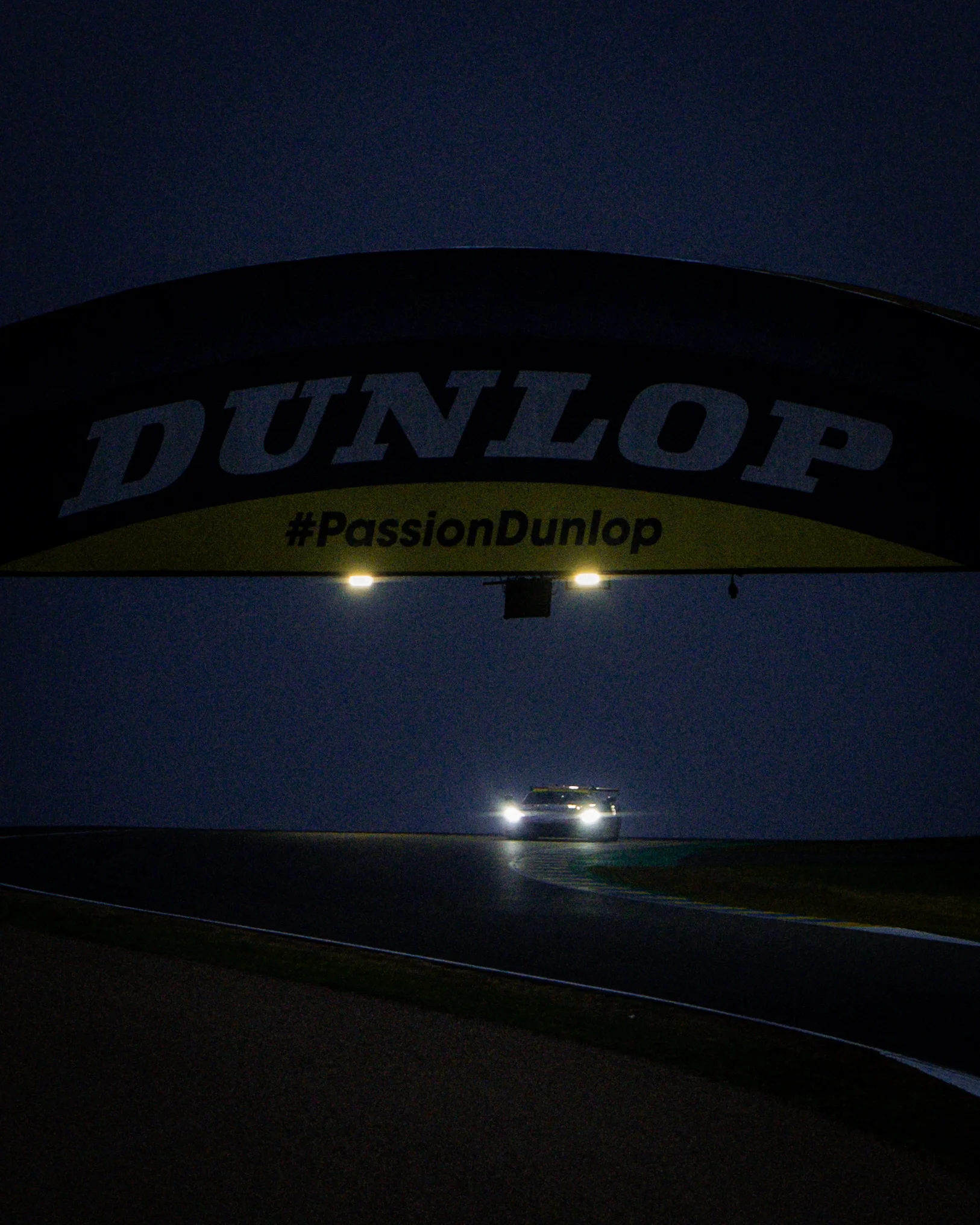 Voiture de course éclairée avec un éclairage puissant, passant sous une arche publicitaire Dunlop lors d'une course de nuit sur un circuit.