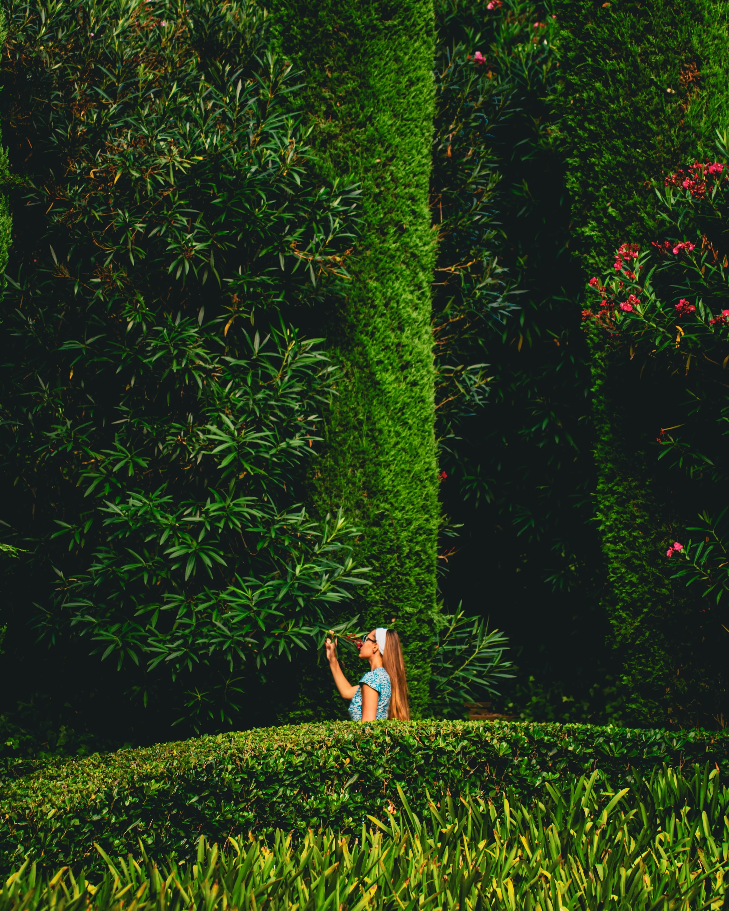 Jeune femme portant des lunettes de soleil, une robe bleue avec des motifs, et un bandeau blanc, se tenant parmi un espace vert dense avec des buissons et un arbre en arrière-plan.