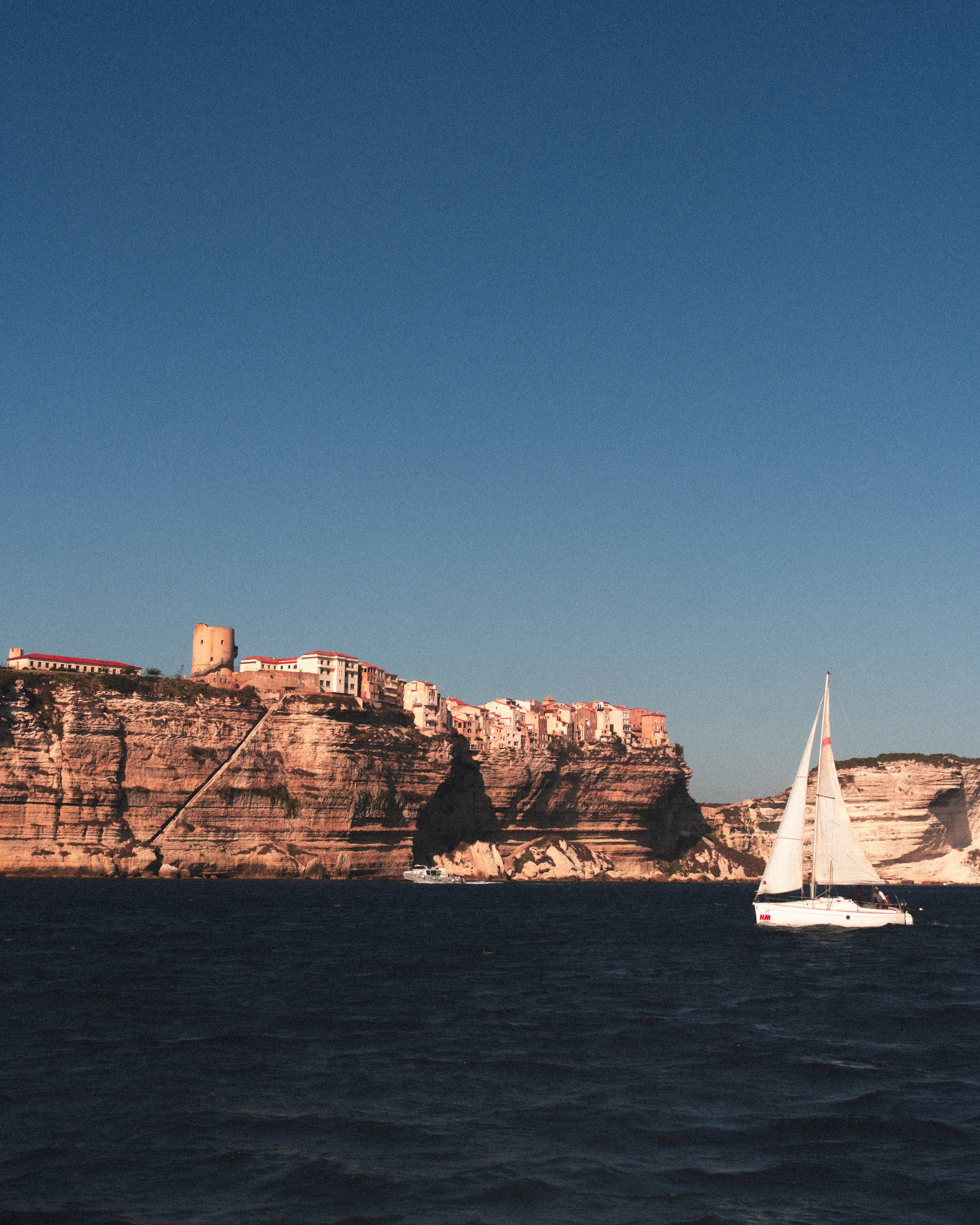 Une côte avec des bâtiments en hauteur et une voile blanche sur la mer.