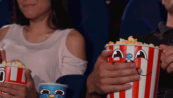 Two people sitting in a movie theater holding large buckets of popcorn, one of them has a smaller drink cup, and they are watching a film.
