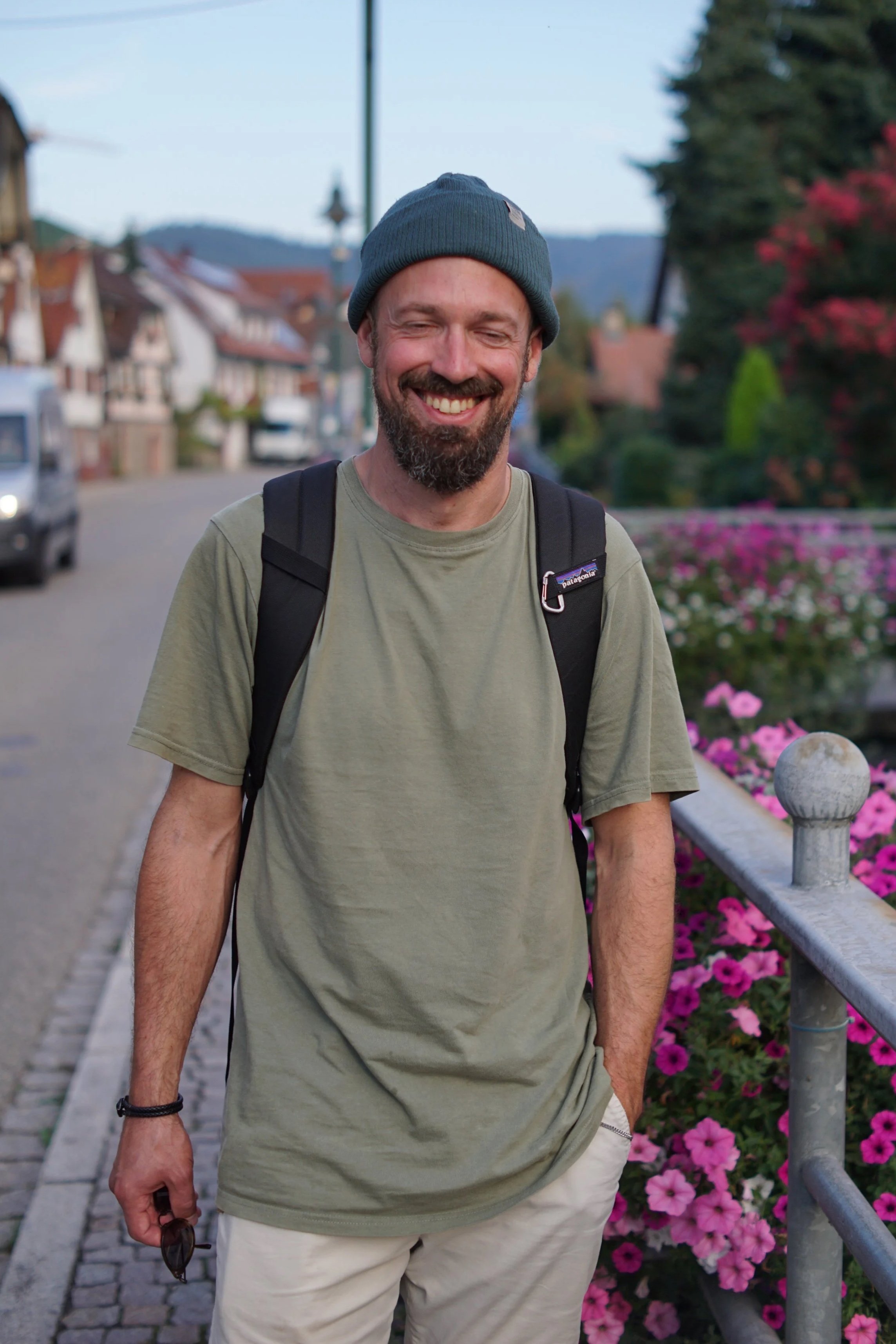 Lächelnder Mann mit Bart, grauem Beanie, grünem T-Shirt und Rucksack steht neben einer Blumenkreation auf einer Brüstung in einer kleinen Stadt.