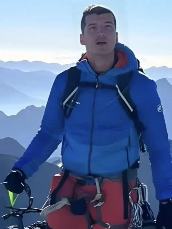 Young man in blue jacket and red climbing gear on mountain summit, with mountain range and sky in background.