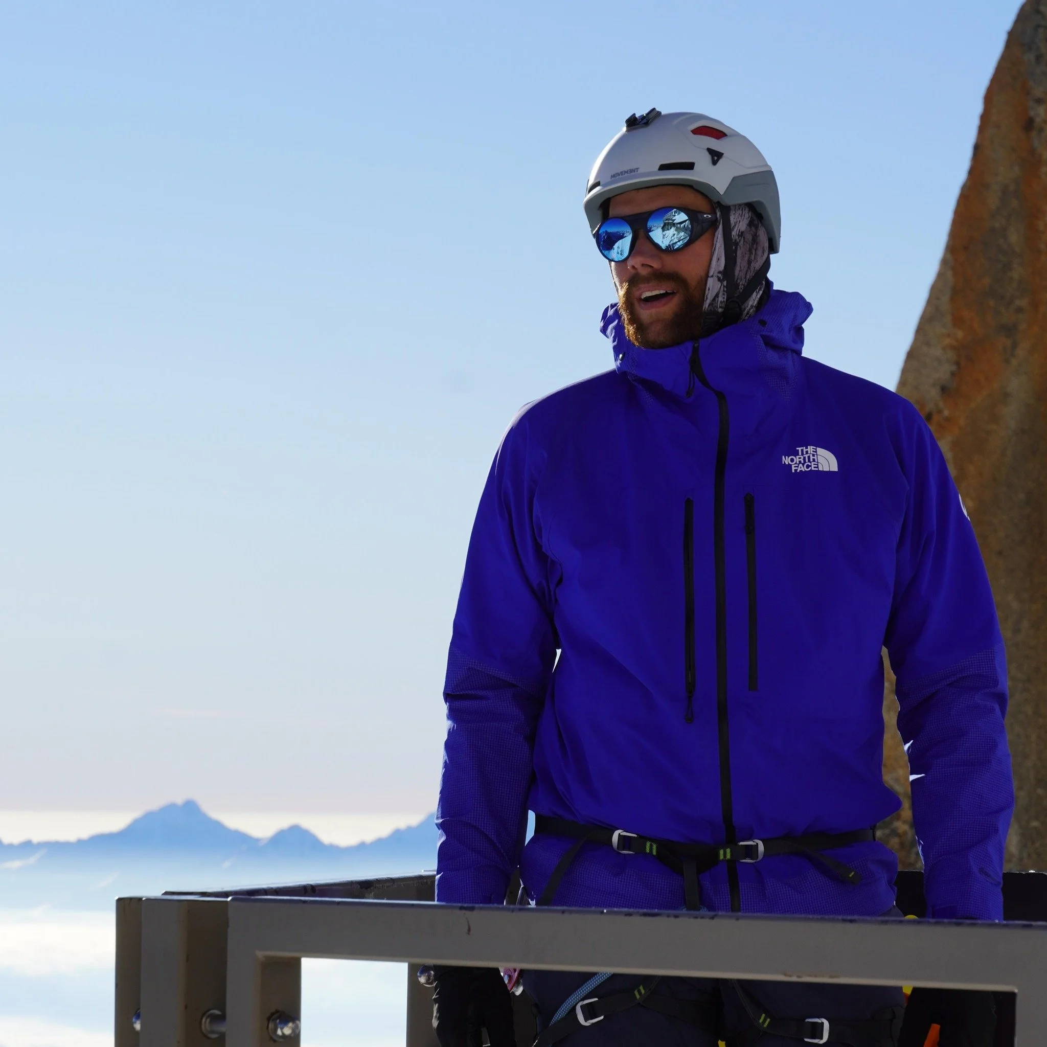 A man dressed in purple outdoor gear, wearing a helmet and sunglasses, stands outdoors with a mountain and sky background.