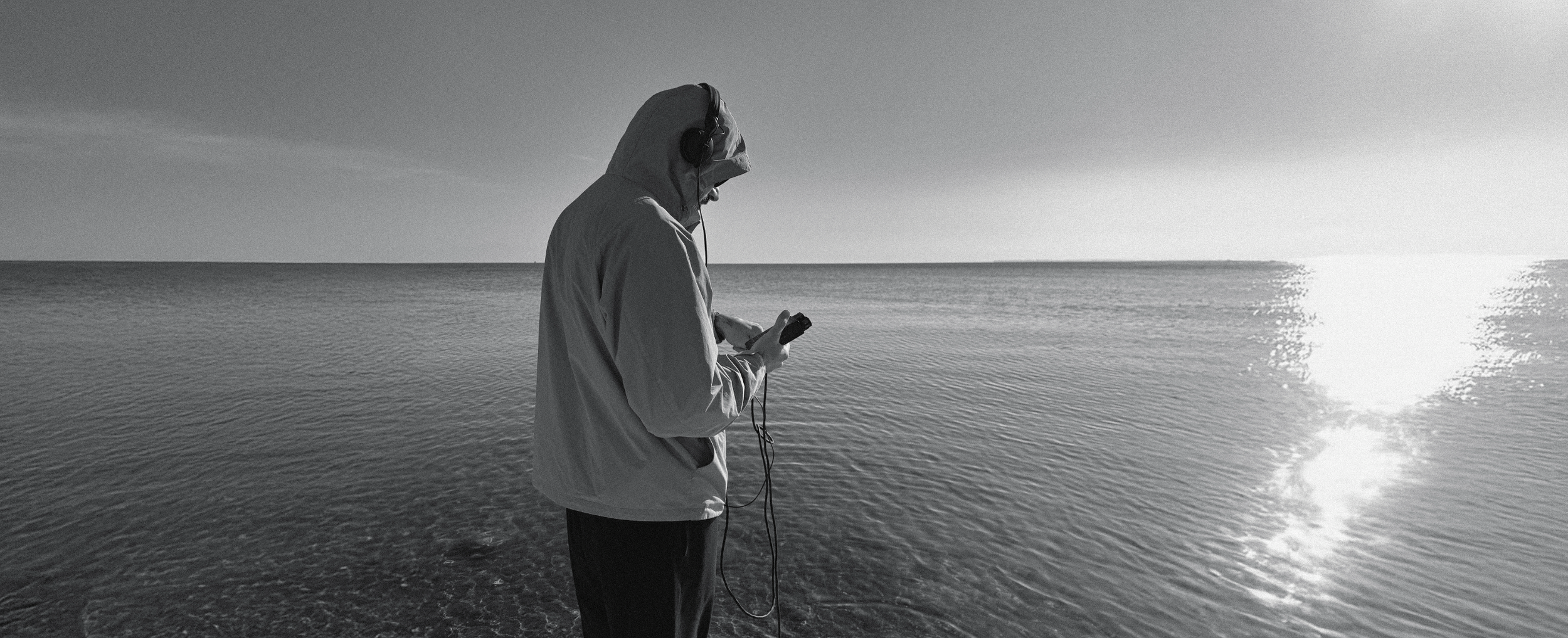 A person wearing a hoodie and headphones, looking at a smartphone while standing at the edge of a body of water during sunset or sunrise.