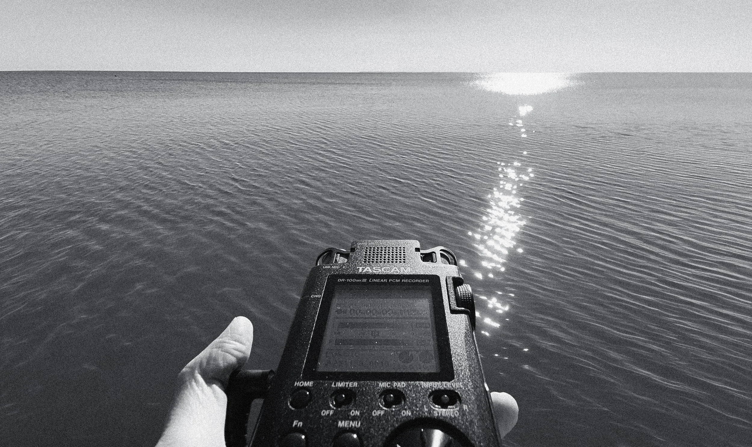 A person holding a portable audio recorder near a calm body of water, with sunlight reflecting on the surface.