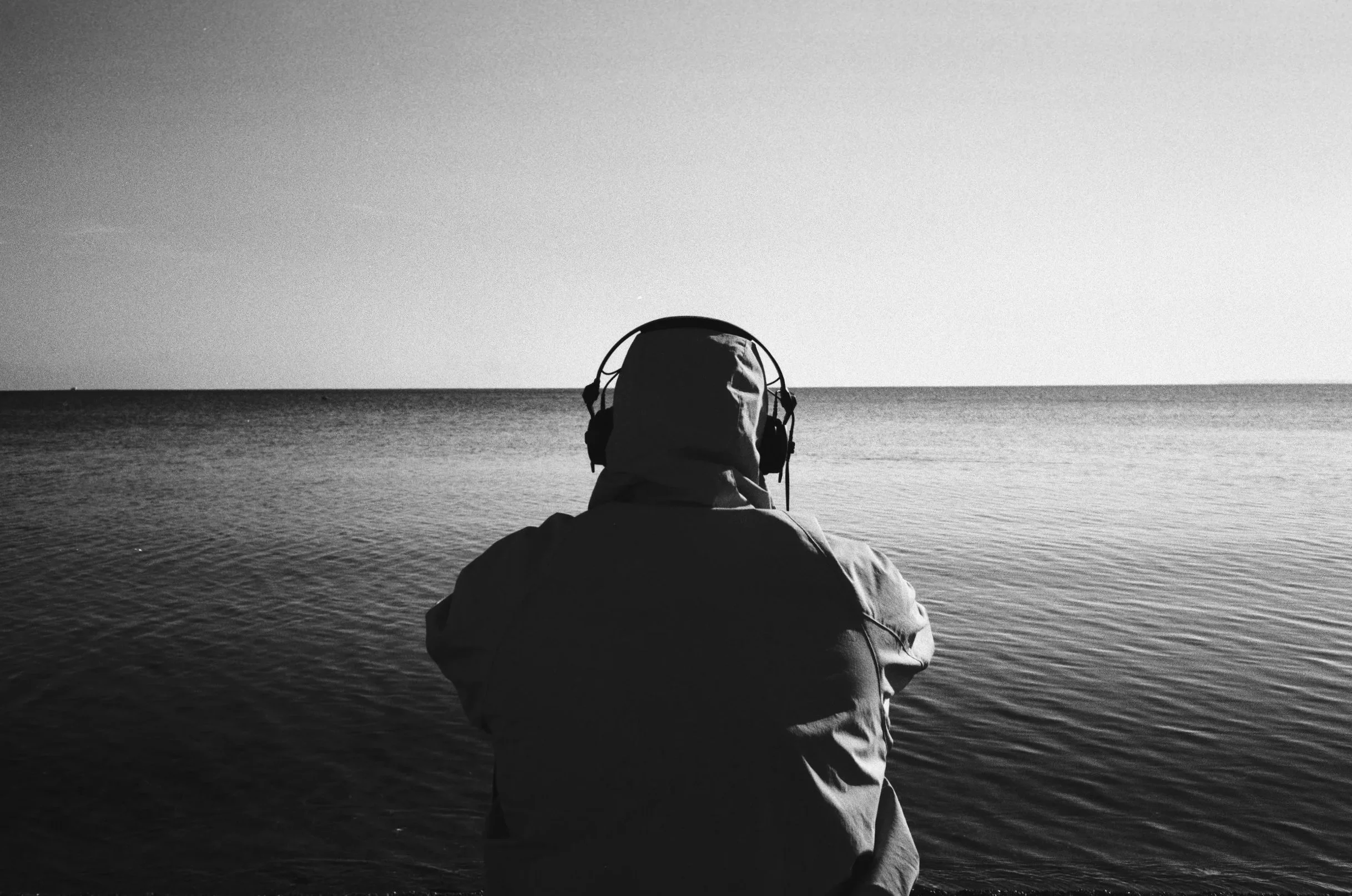 A person with a hood and headphones sitting by the water, facing the horizon on a black-and-white photo.