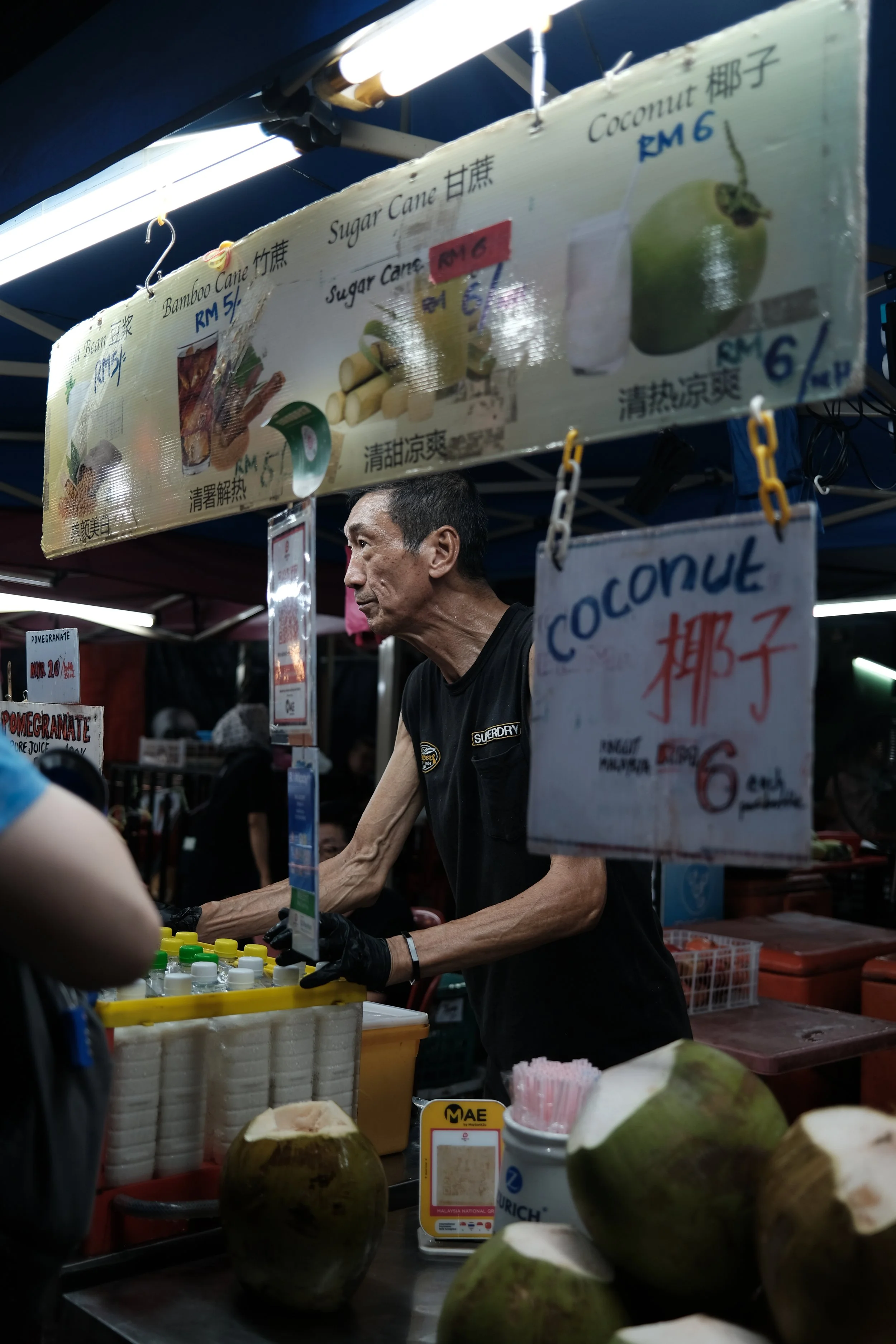 A street vendor preparing fresh coconuts at night market stall with signs listing coconut and sugar cane drinks. Coconuts are on the counter, and the vendor is wearing a black shirt, with other market stalls visible in the background.