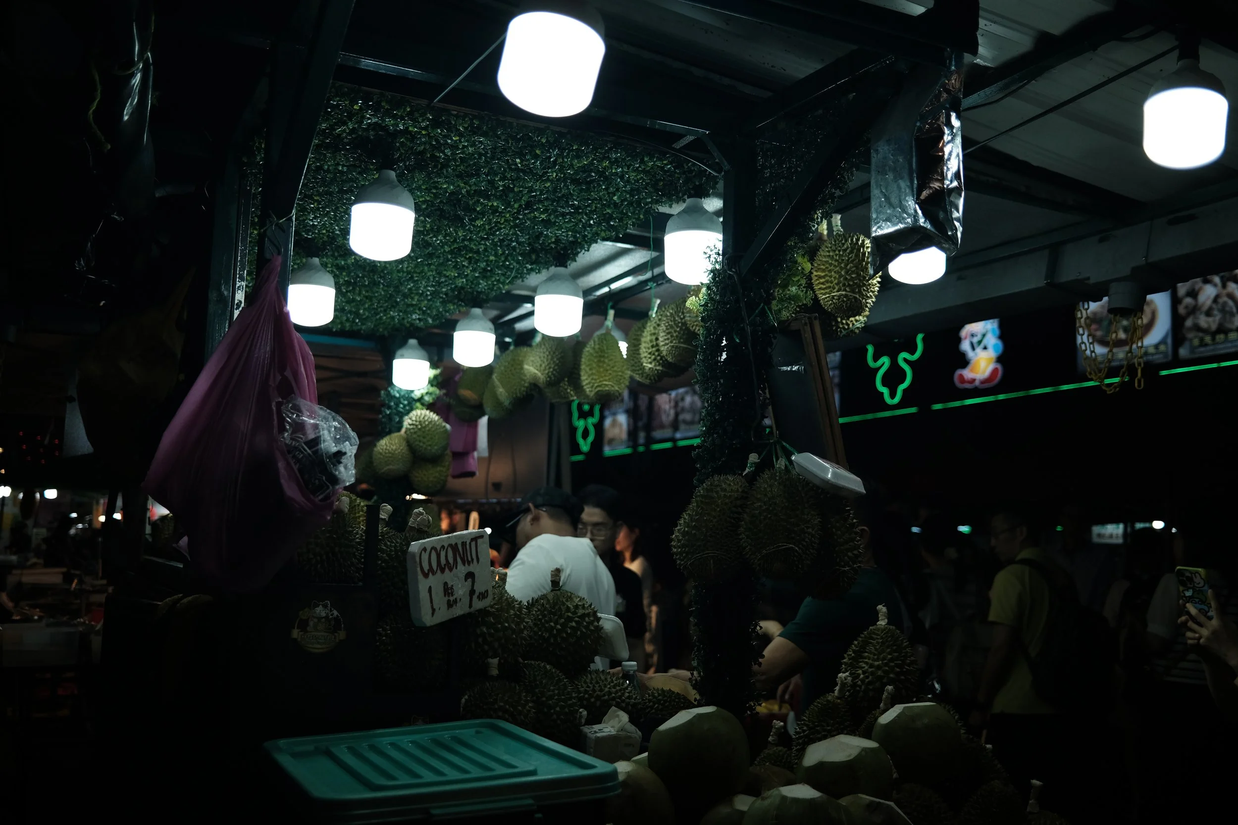A street market stall selling durian fruit, with hanging durians and a sign indicating the price of coconut, under bright overhead lights and surrounded by people.