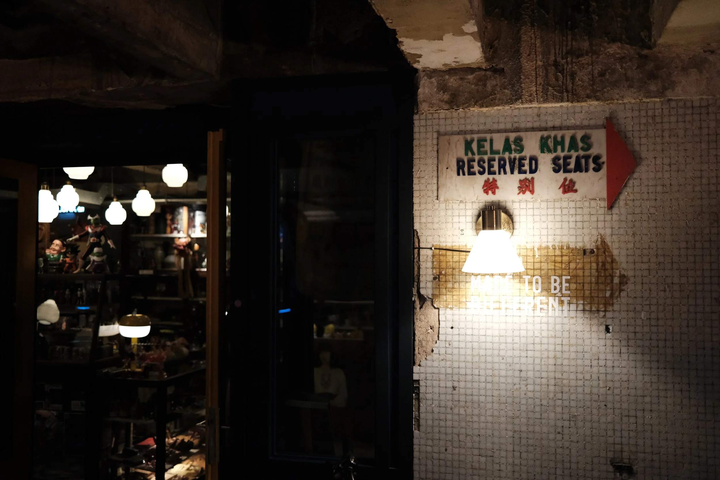 A sign in English and Chinese indicating reserved seats at a restaurant entrance, with another sign below stating 'HOW TO BE ELEGANT,' illuminated by a wall-mounted light fixture, with a dark interior space visible in the background.