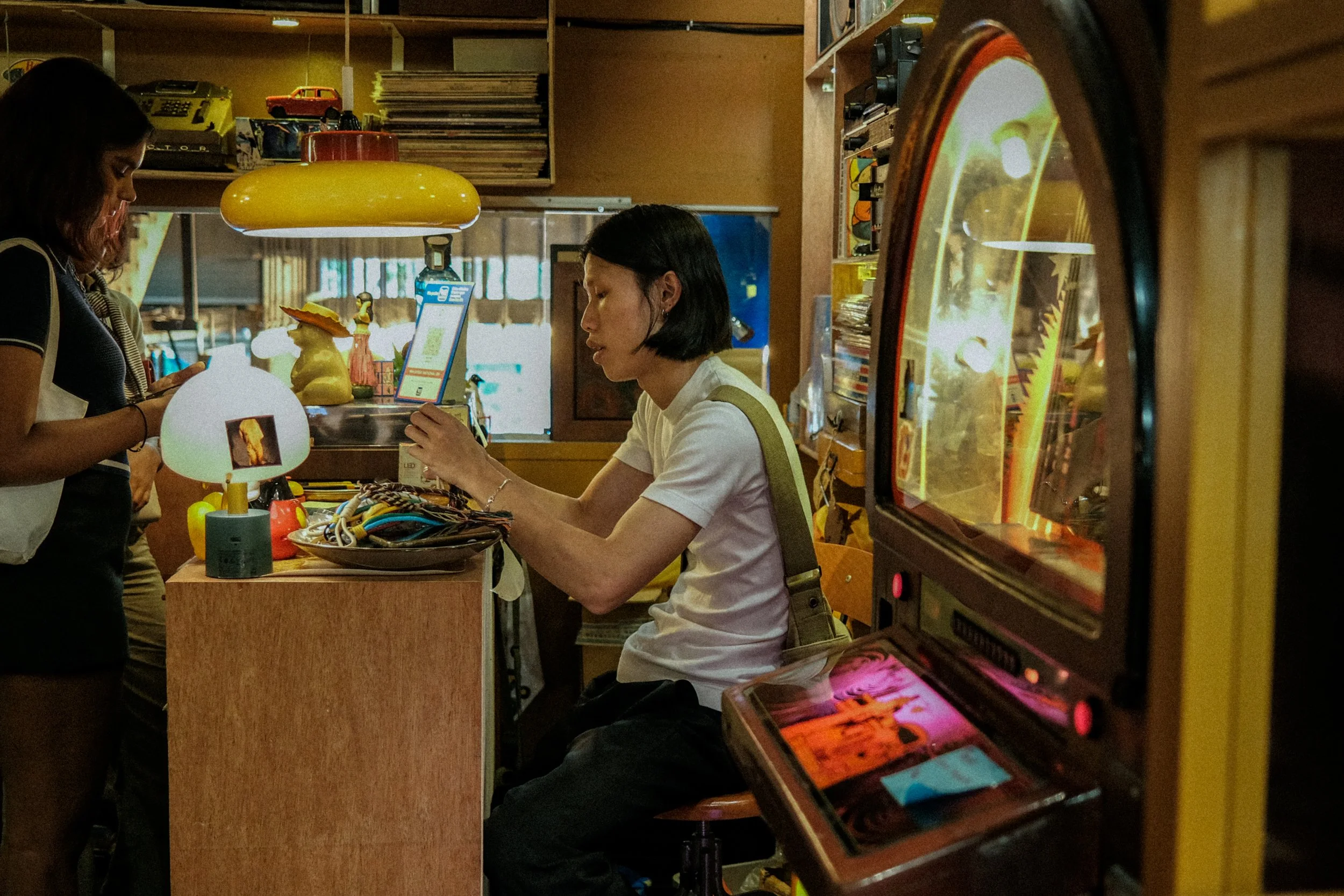 A woman with black hair sitting at a counter in a vintage arcade, using a touchscreen device, with a woman standing next to her looking at her phone, surrounded by retro decor and arcade machines.