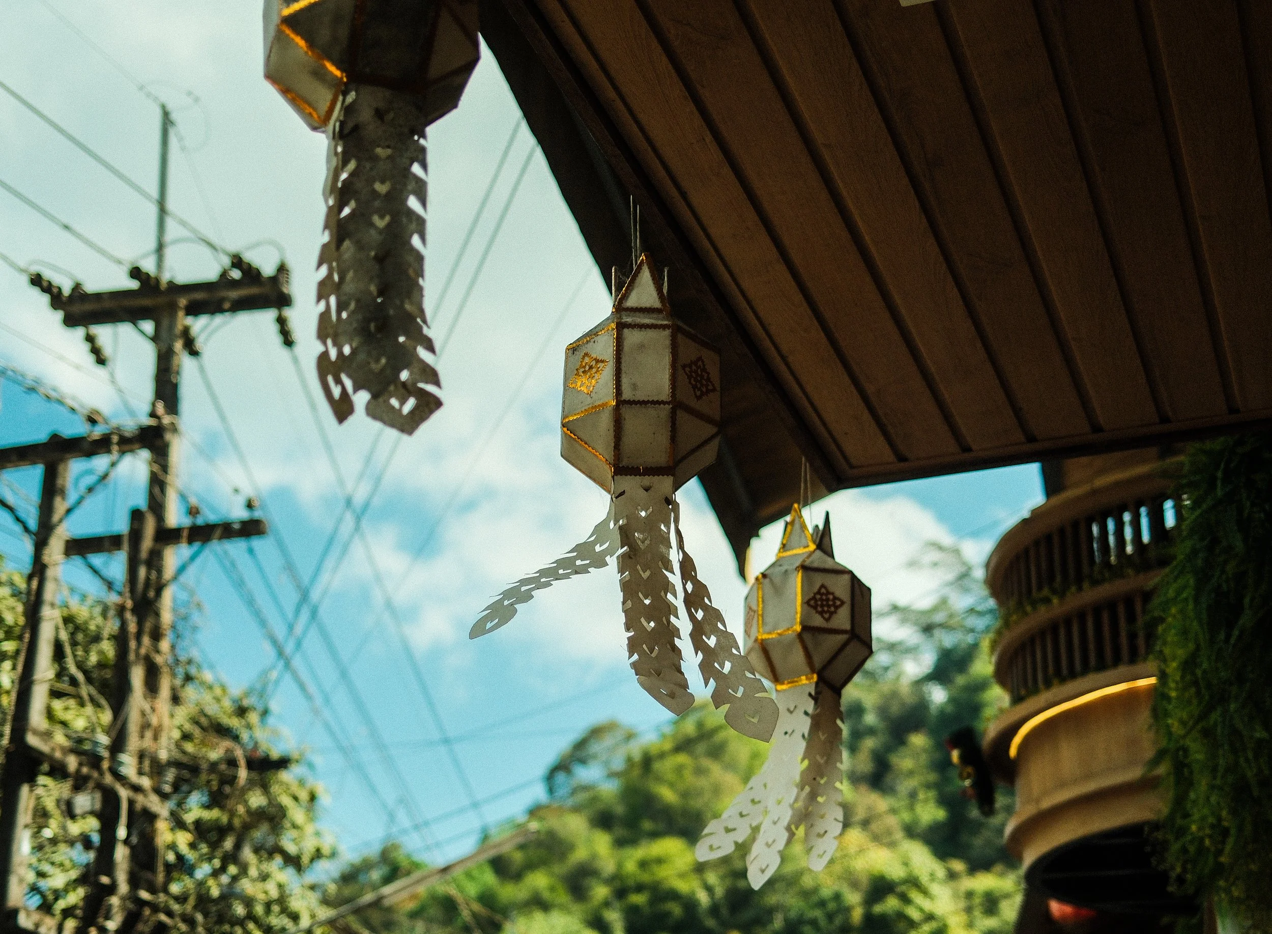 Three paper lanterns hanging from a wooden ceiling outdoors with utility poles and clear blue sky in the background.