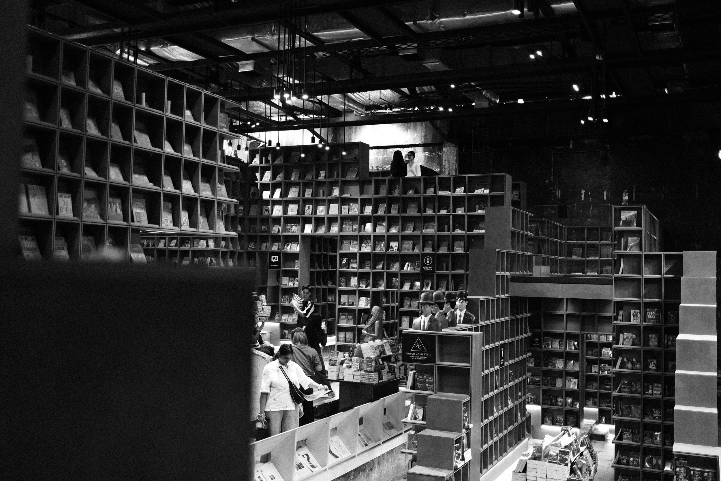 Black and white photo of a bookstore with tall shelves filled with books, customers browsing, and a counter with mannequins wearing hats.