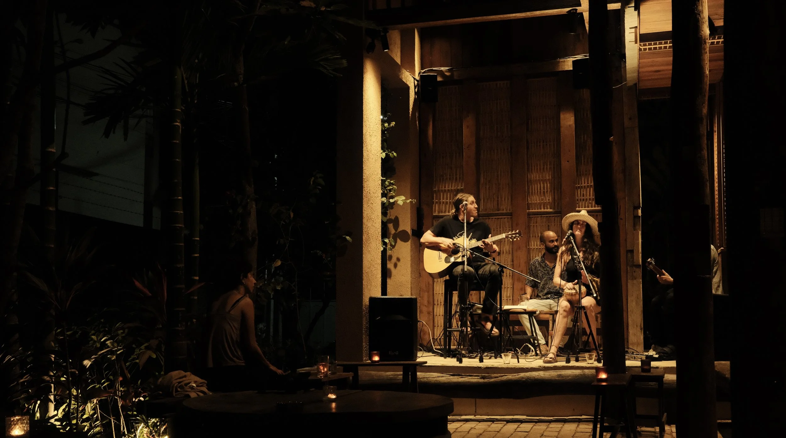 Musicians performing on a small stage at night, with a man playing guitar, a woman singing into a microphone wearing a large hat, and a man in the background playing drums. Small candles and drinks are on tables in the foreground.