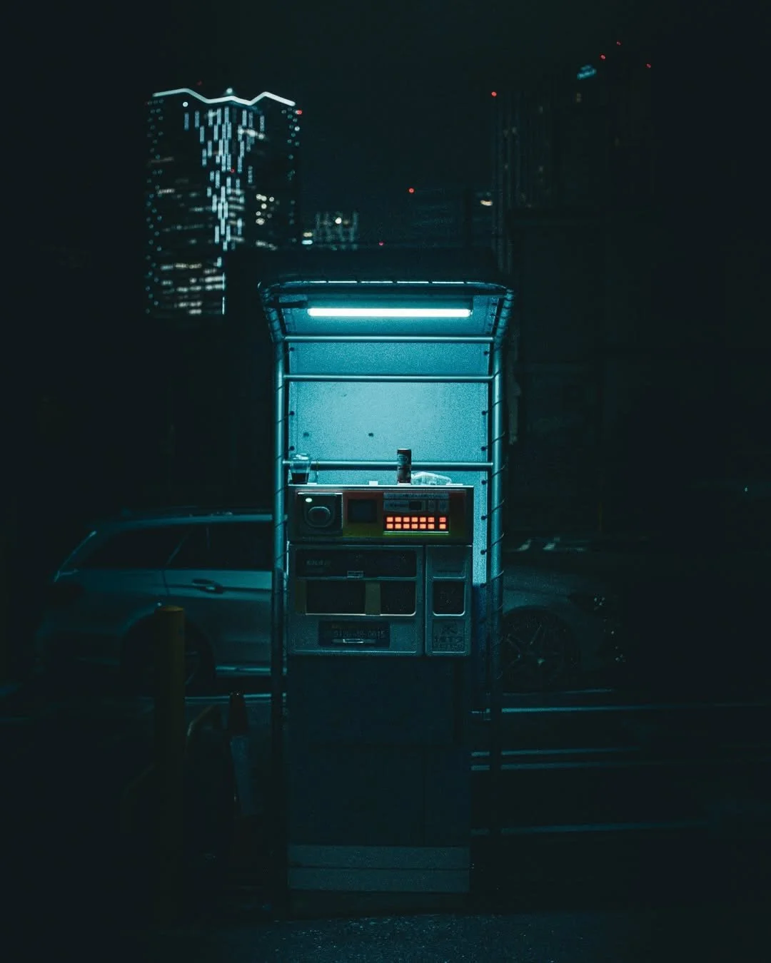 A nighttime city scene with a vending machine and a parked car illuminated by blue neon light. Tall buildings with lit windows are visible in the background.