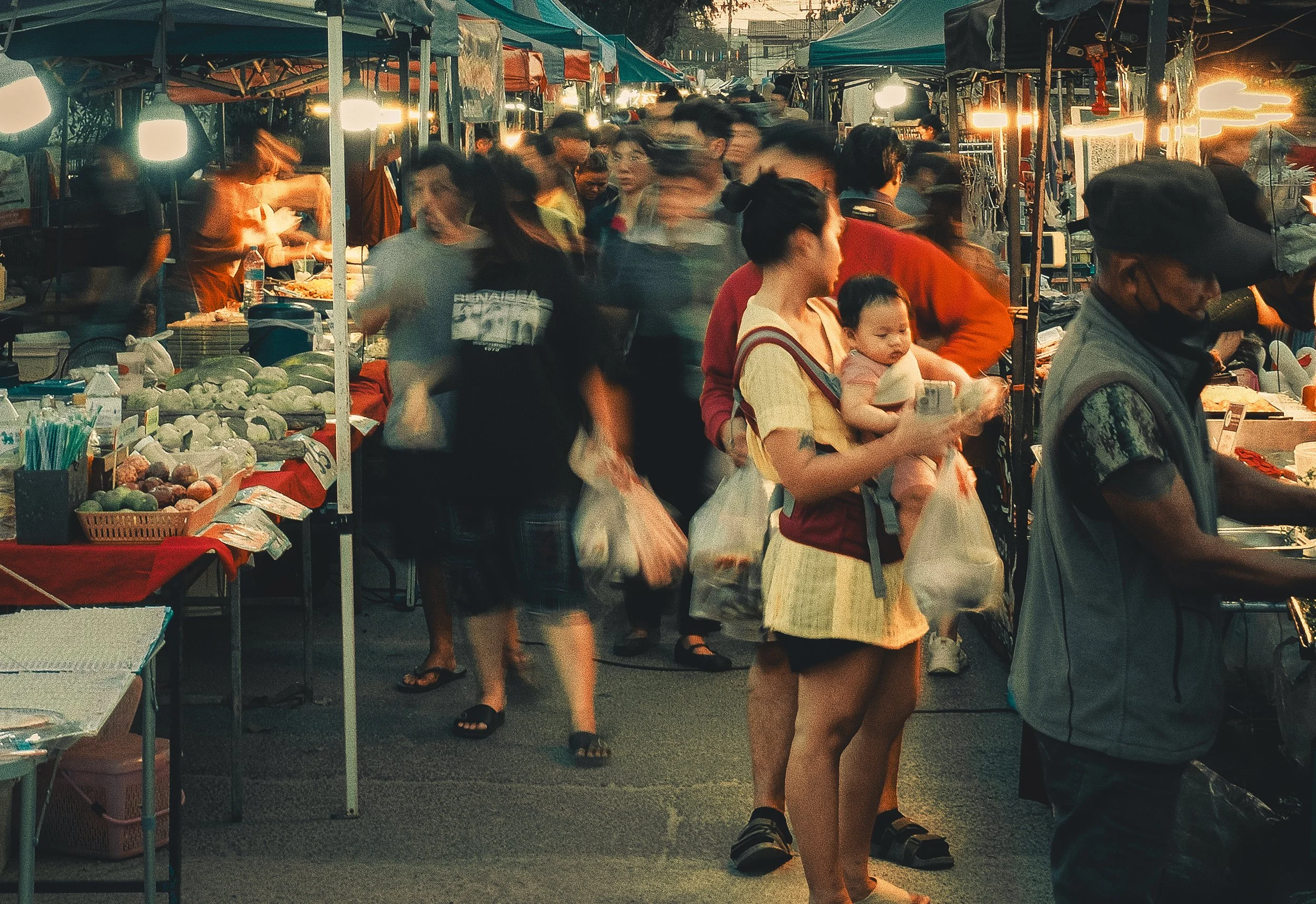 Crowded outdoor night market with vendors and shoppers, some carrying bags, stalls offering produce, and blurred lights and people indicating movement.