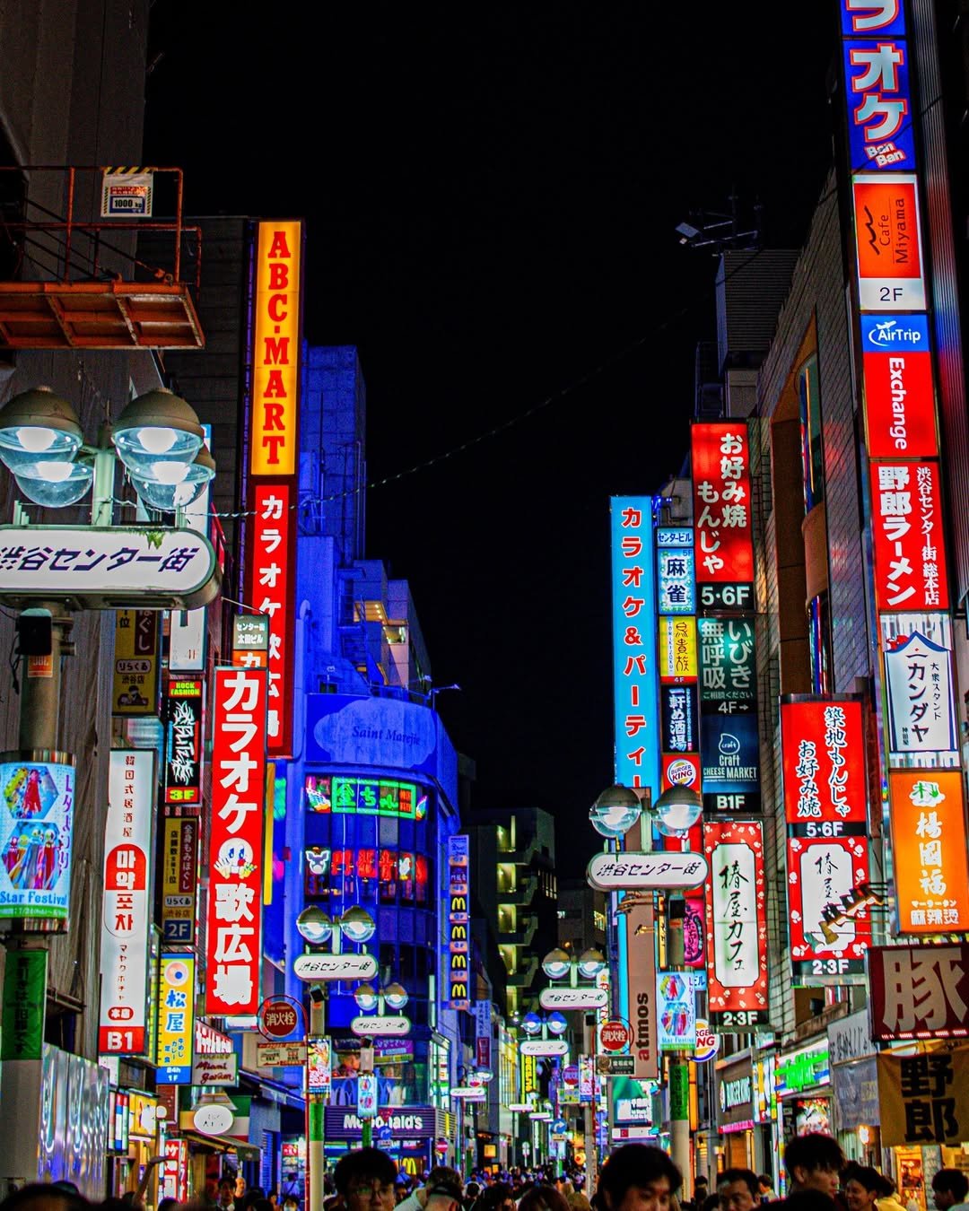 Nighttime street scene in Osaka, Japan, with bright neon signs in Japanese and English advertising various shops, restaurants, and entertainment venues, crowded with pedestrians.