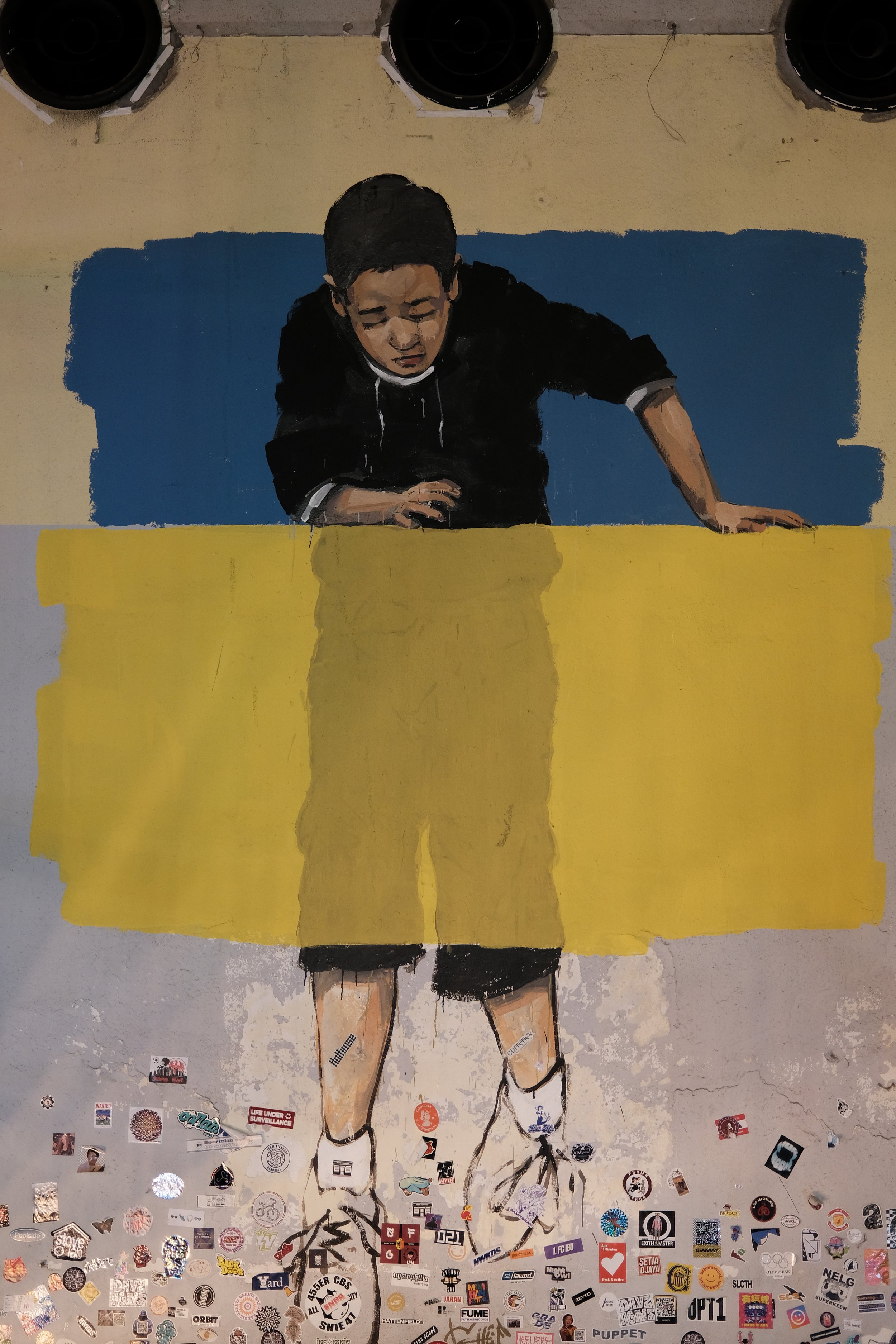 A mural of a young person with glasses, looking down, with hands on a yellow table, painted with dark hair, black hoodie, and shorts, amid stickers on the wall below.