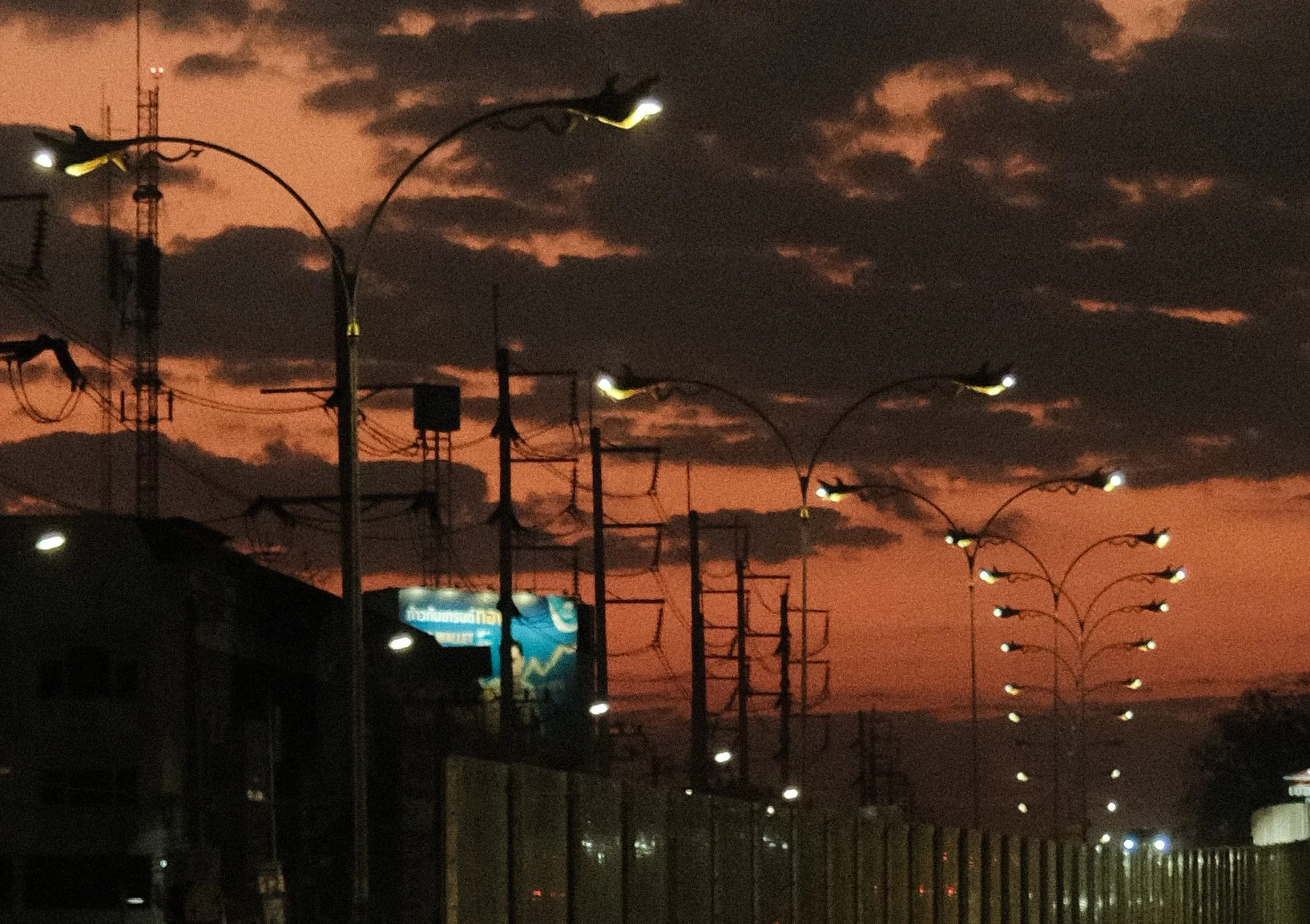 City street during sunset with dark clouds, illuminated streetlights, and power lines.