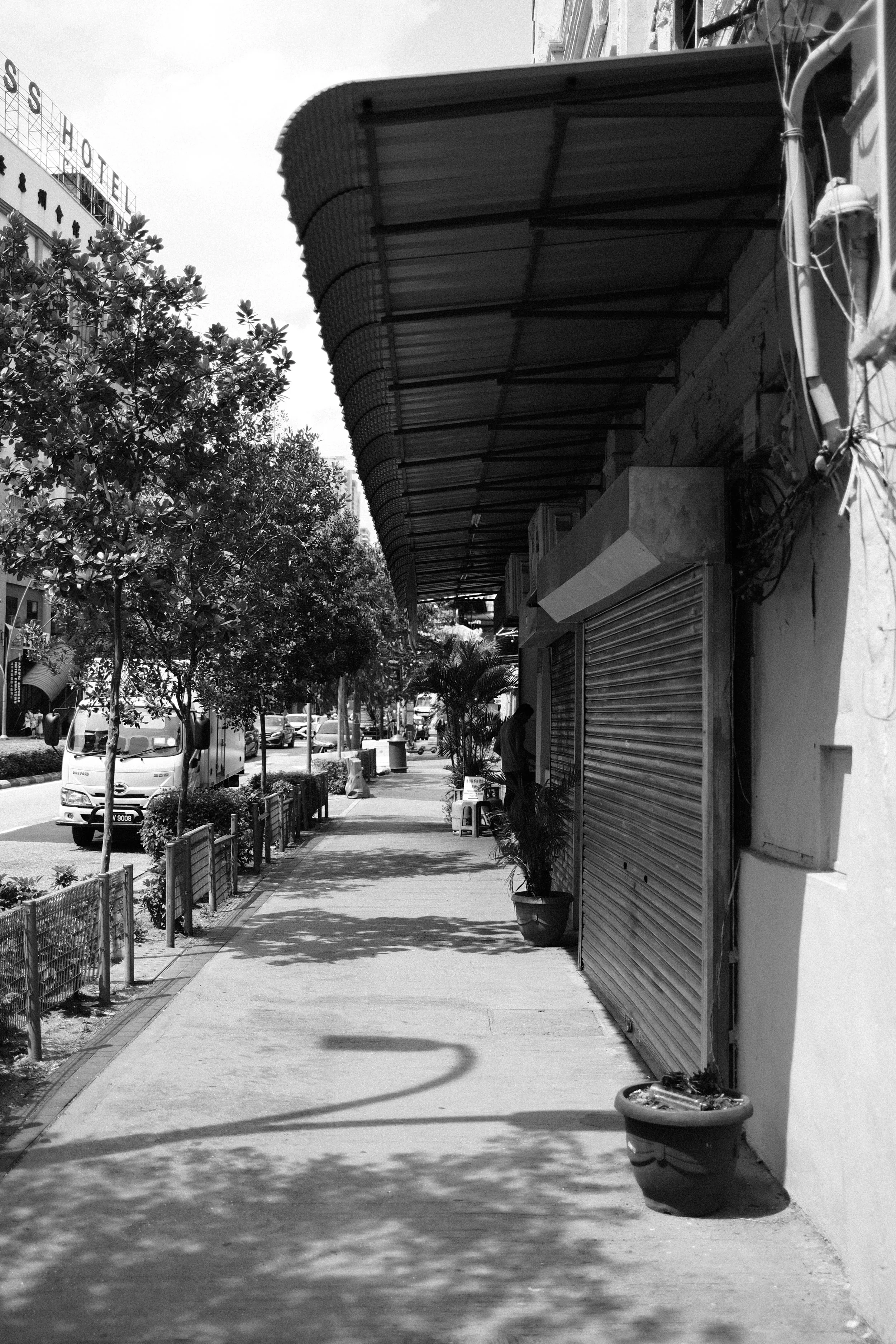 Empty sidewalk with closed storefronts, potted plants, and trees, casting shadows, in an urban area.