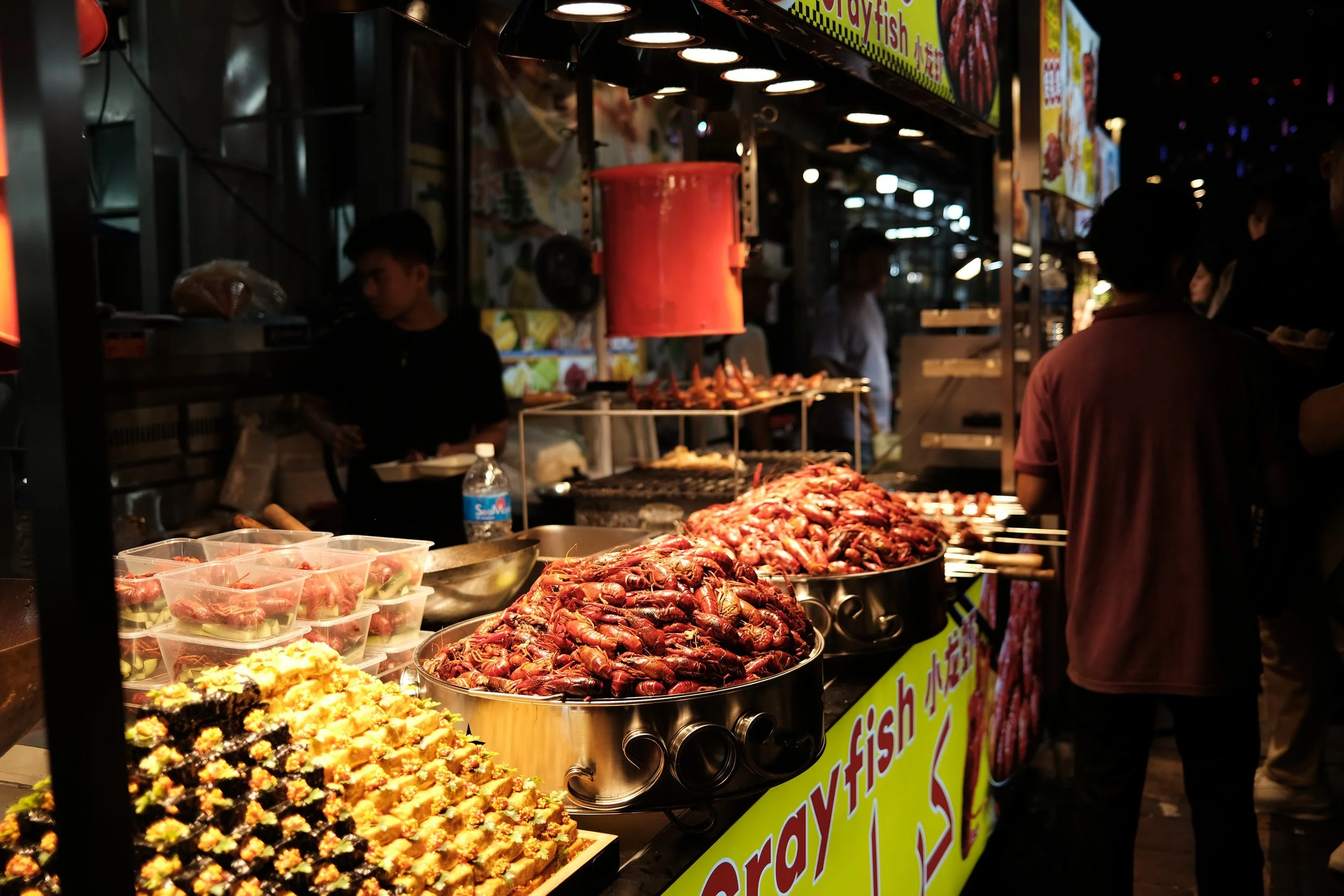 Night market seafood stall with cooked crawfish, sushi, and skewered seafood, surrounded by people