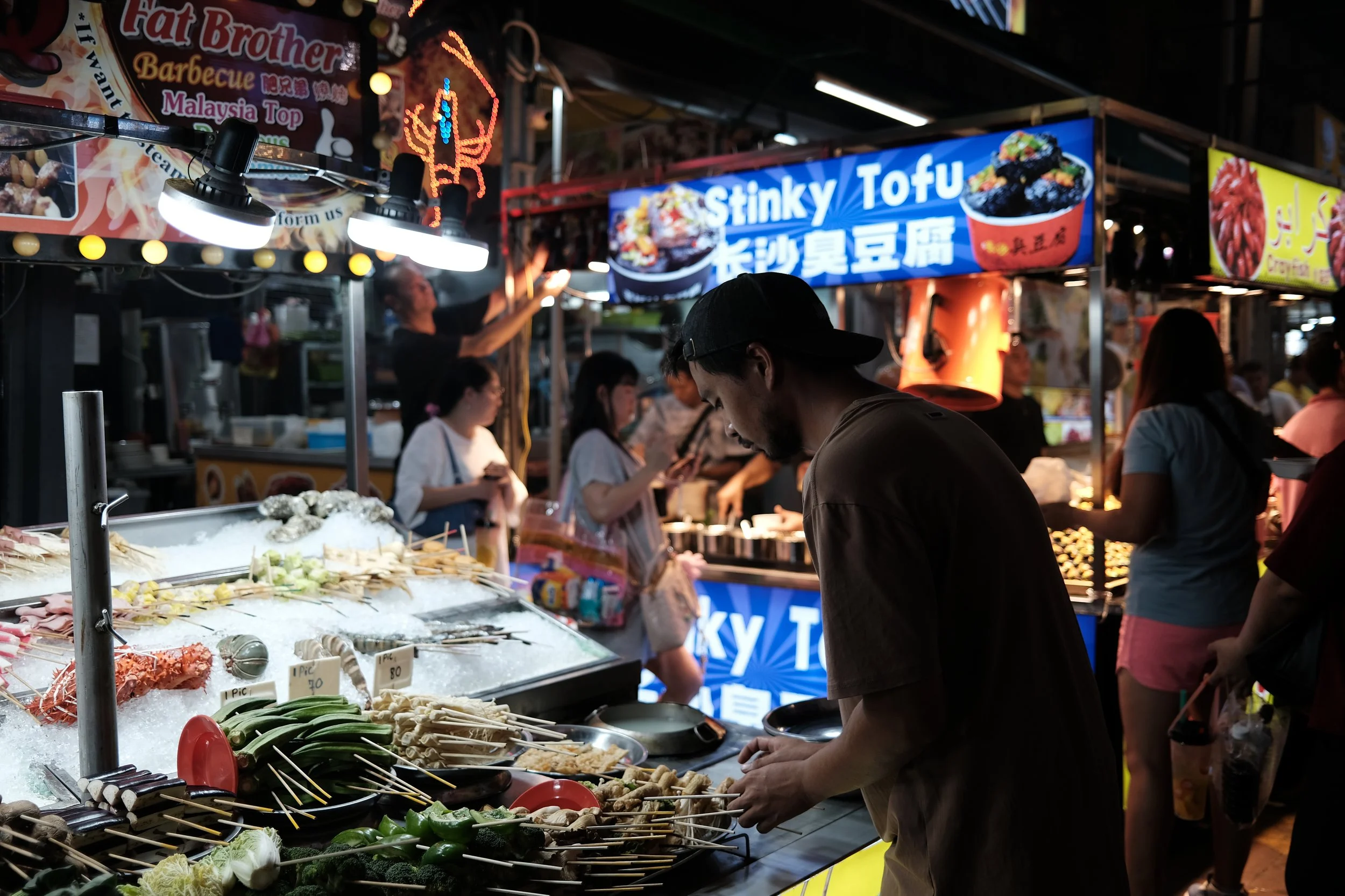 Night market street food stall with skewered vegetables, seafood, and meats, illuminated by bright lights, with people shopping and preparing food, and signs advertising dishes like stinky tofu.