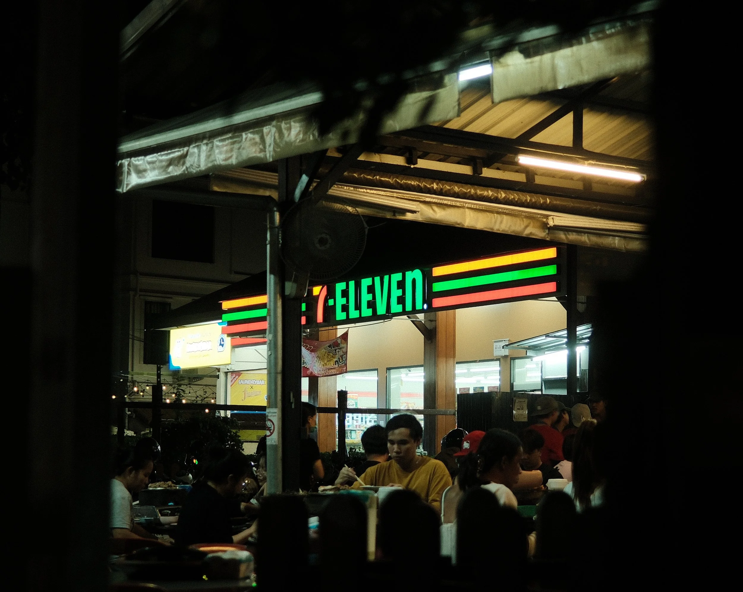 Night view of a 7-Eleven convenience store with illuminated sign and people inside.