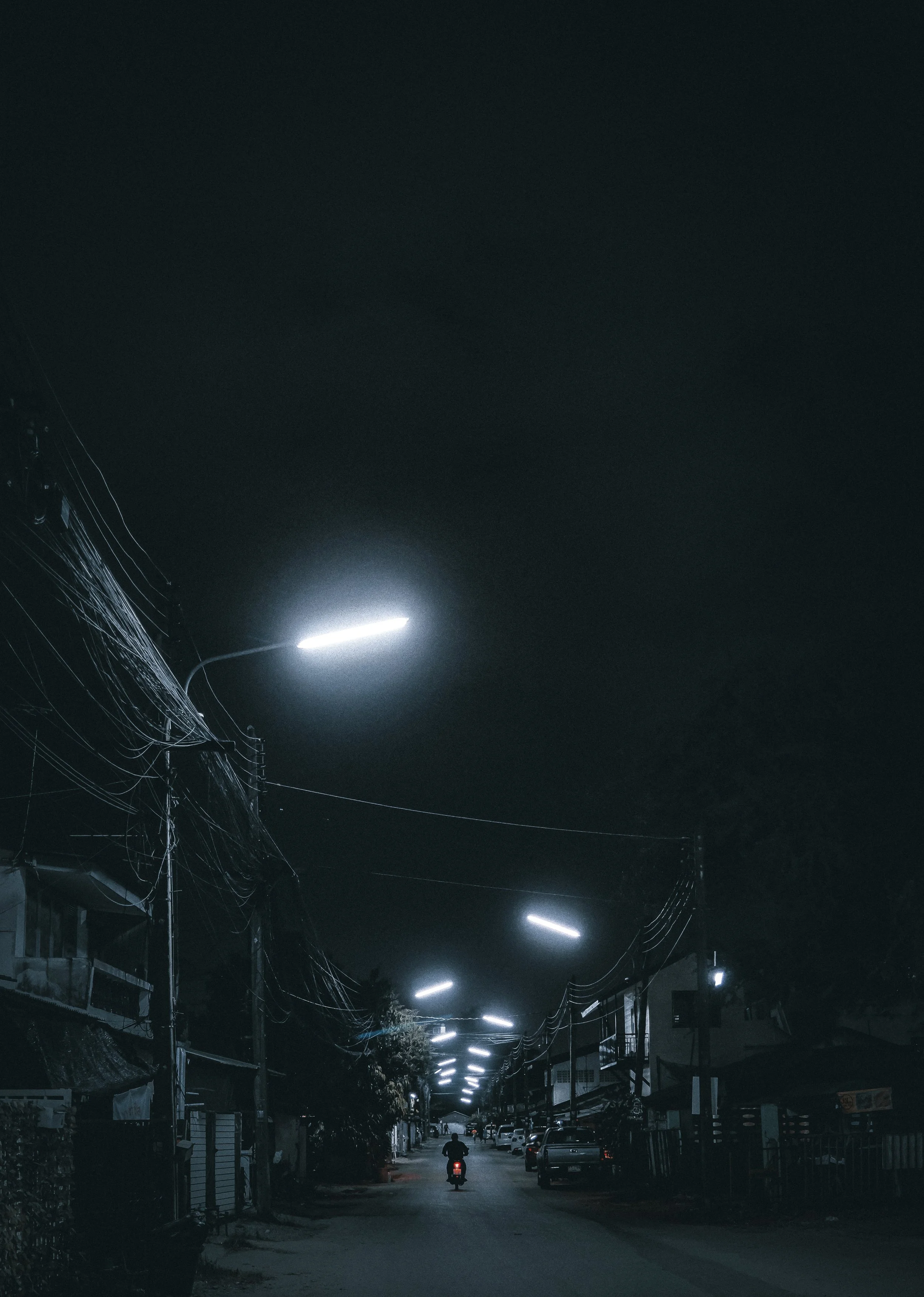 A person riding a motorcycle down a dark street illuminated by streetlights at night, with houses and parked cars on either side.