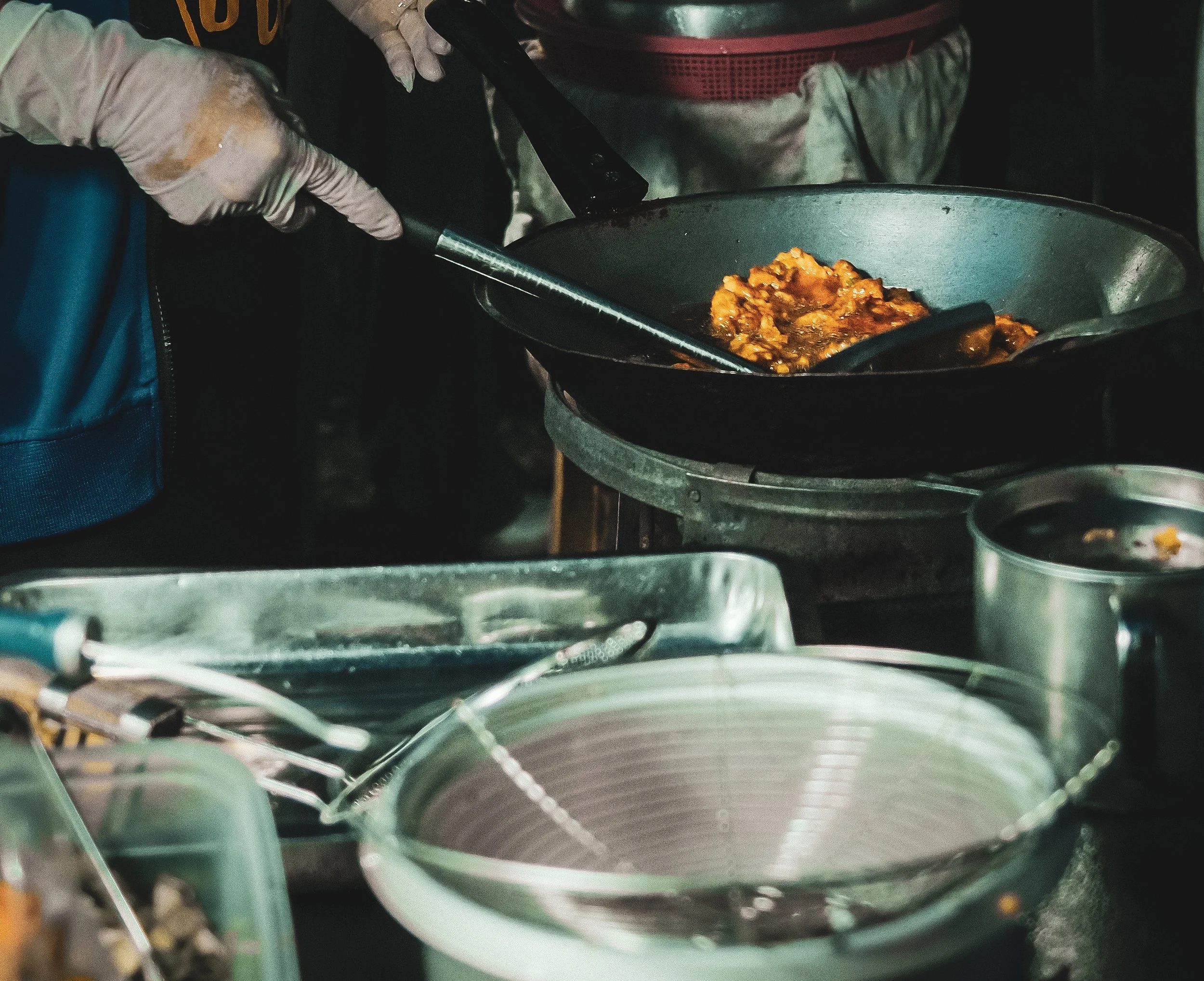 A person cooking stirring a dish in a black skillet over a stove, wearing striped pants and gloves, with various kitchen utensils and containers visible around.
